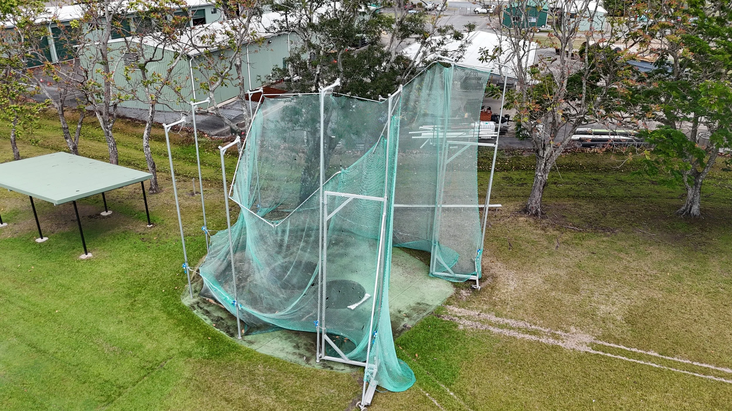 An outdoor batting cage with a green net and white frame, located in a grassy area with trees. A green roofed building and parking lot are visible in the background.