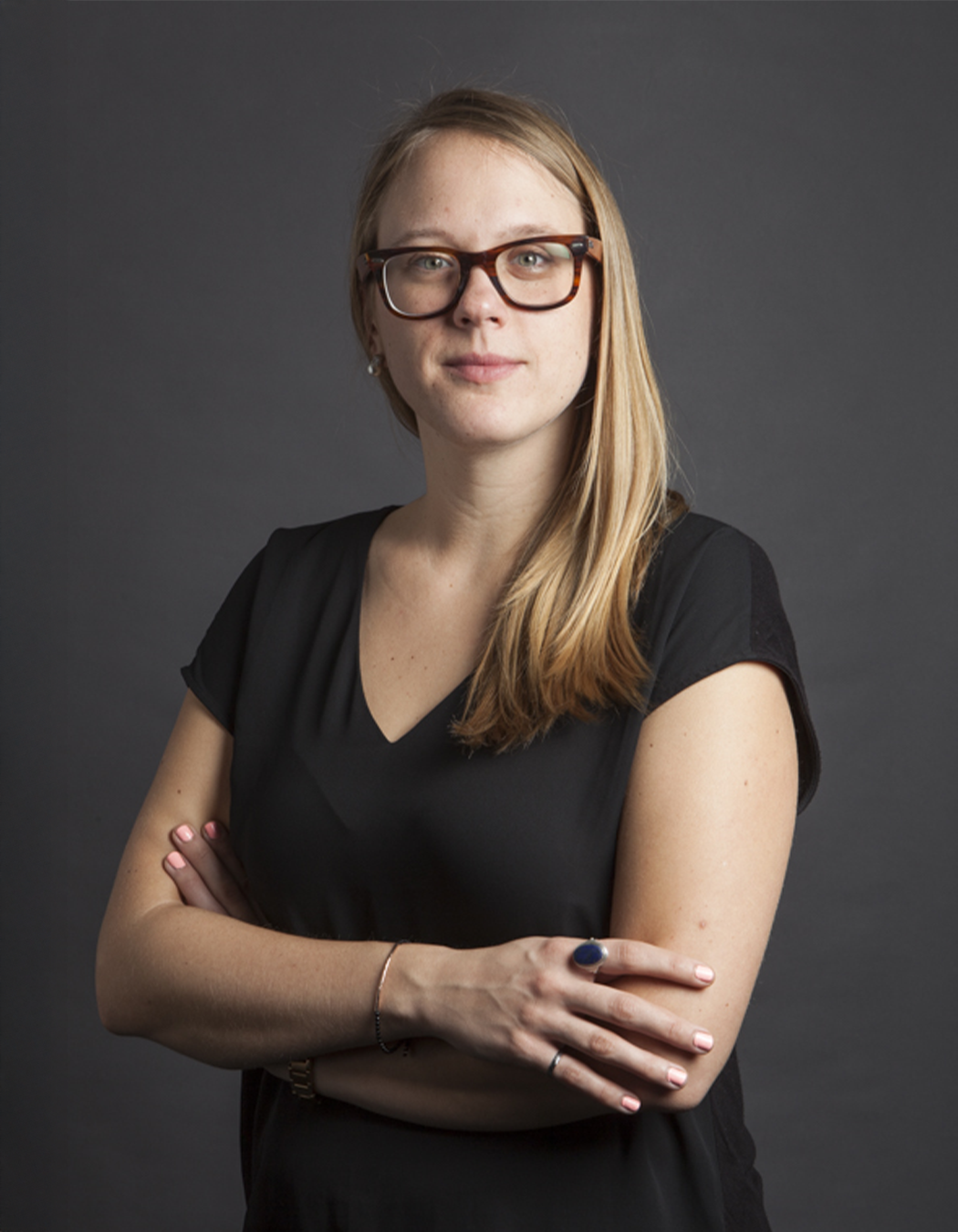 A woman with long blonde hair, wearing glasses and a black top, posing with crossed arms against a dark gray background.