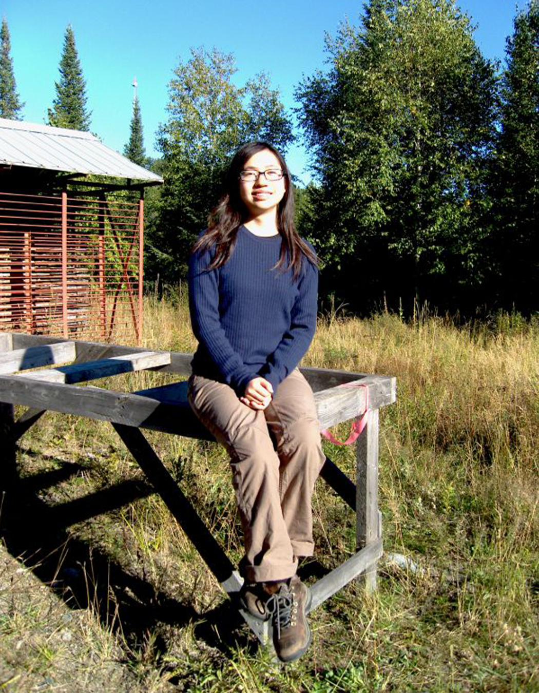 Young woman with glasses sitting on a wooden picnic table outdoors, surrounded by tall grass and trees under a clear blue sky.