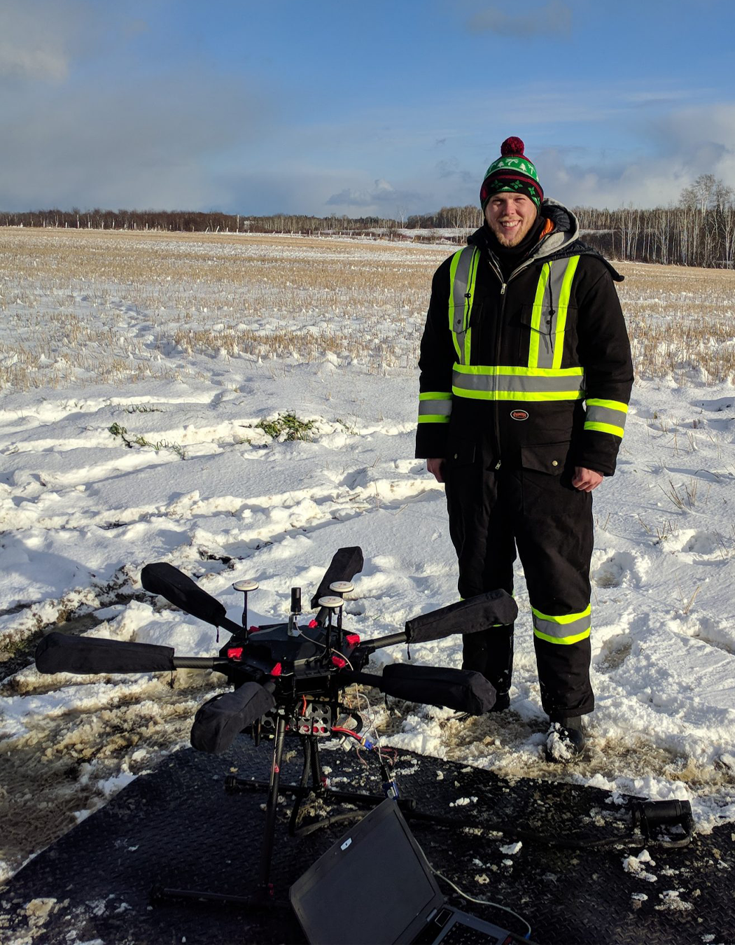 A man dressed in a black jacket with reflective yellow stripes, standing in a snowy field with a drone and a laptop in front of him, under a partly cloudy sky.