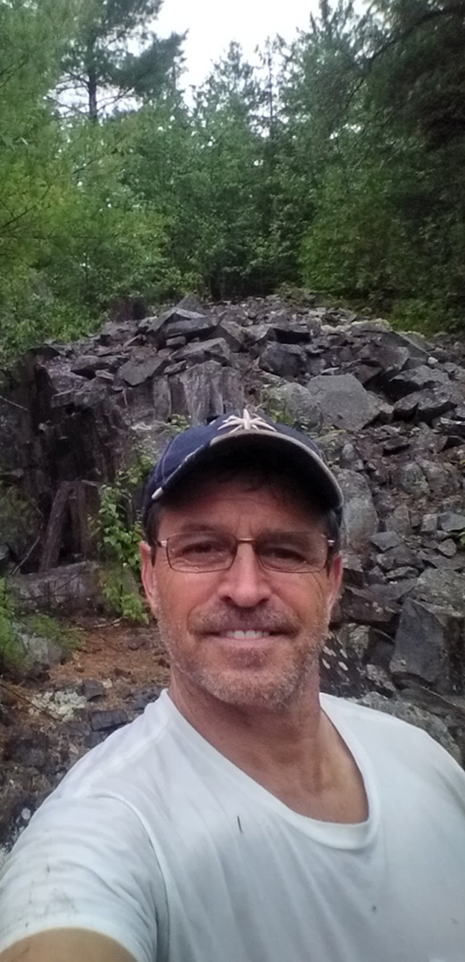 A man taking a selfie outdoors in front of a rocky hillside with green trees and foliage.