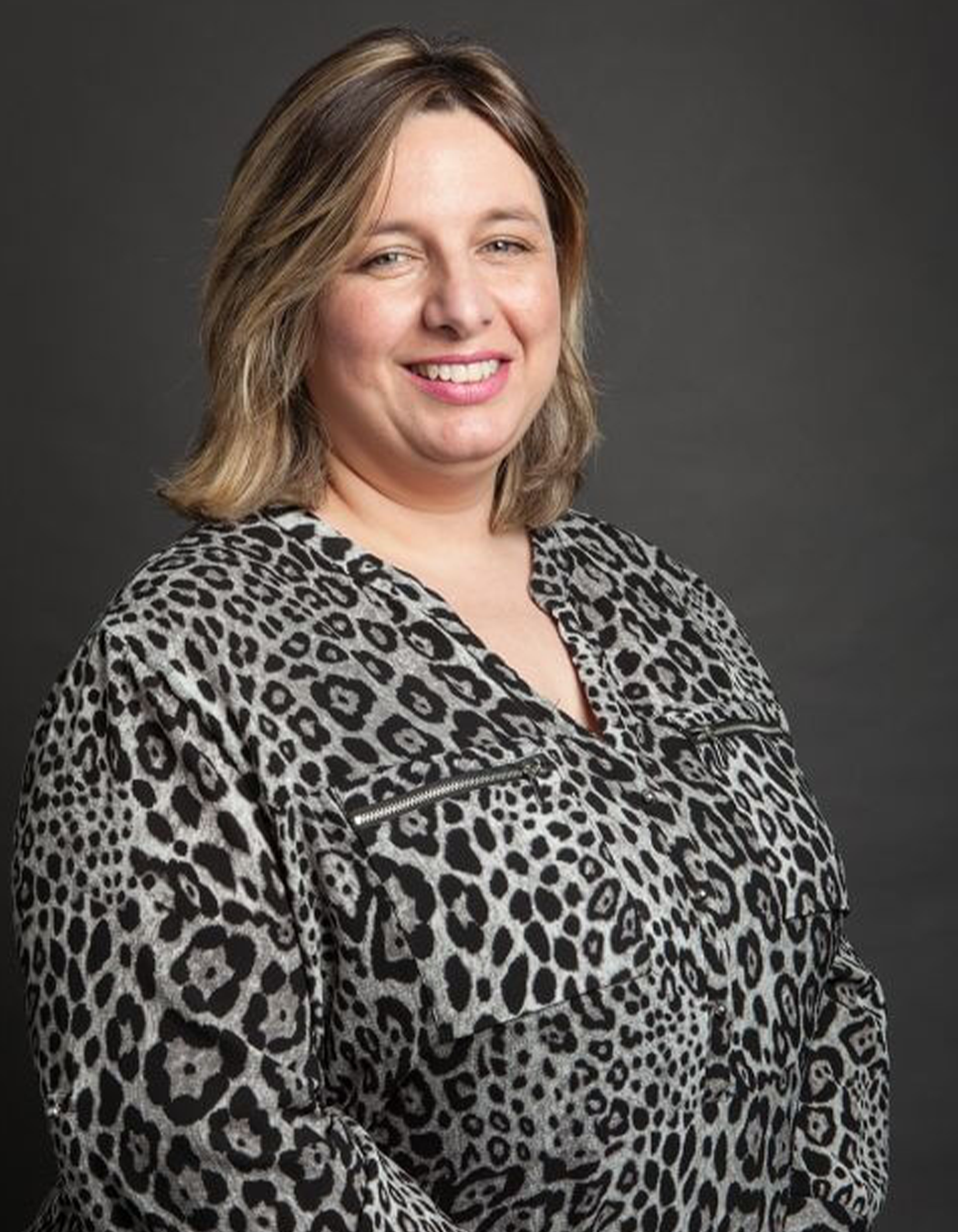 A woman with shoulder-length light brown hair and a smile, wearing a black and white leopard print blouse, posing against a dark gray background.