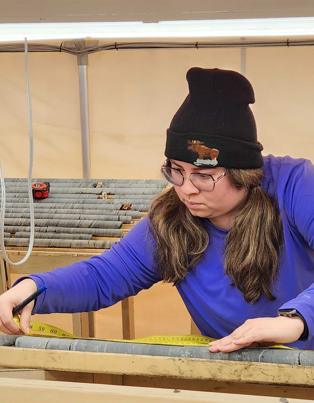 A woman with long brown hair, wearing glasses, a black beanie, and a blue long-sleeve shirt, is logging core with a yellow measuring tape inside a workspace or workshop.