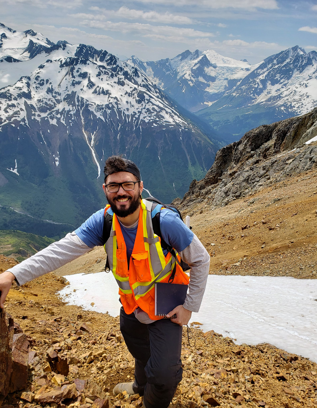 Hiker in a blue shirt and orange safety vest smiling on a rocky mountain trail, with snow-capped mountains and greenery in the background.