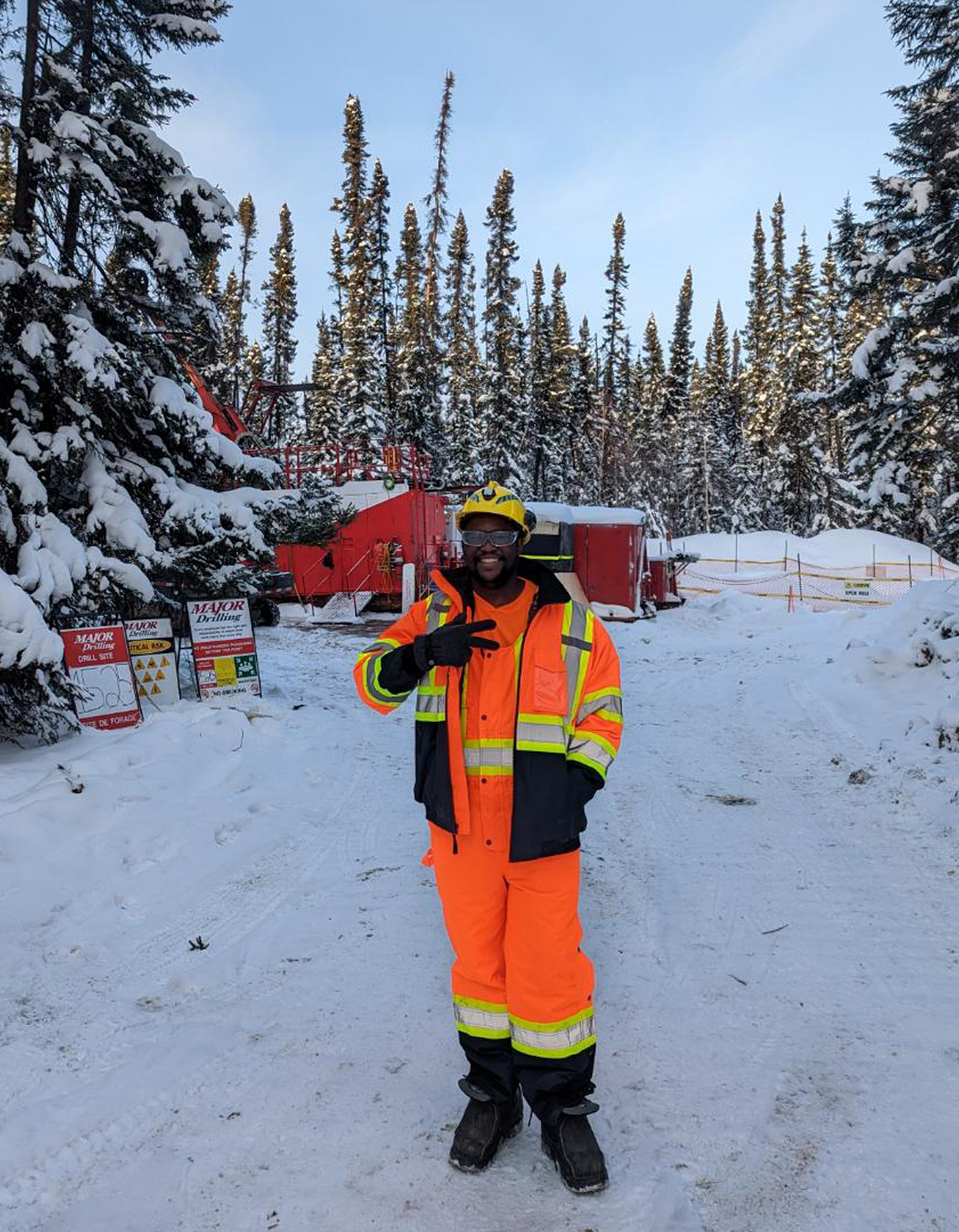 A person in a high-visibility orange and black safety suit and helmet, standing on snow-covered ground in front of snow-laden trees and red drilling equipment, at a drilling site in a snowy forest.
