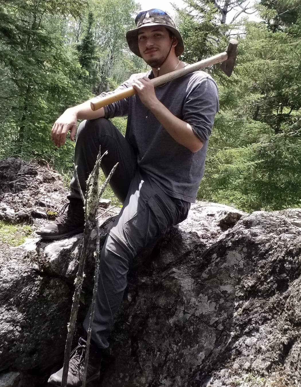 A man in outdoor hiking gear, wearing a hat and sunglasses, sitting on a large rock in a forested area, holding a geotul resting on his shoulder.
