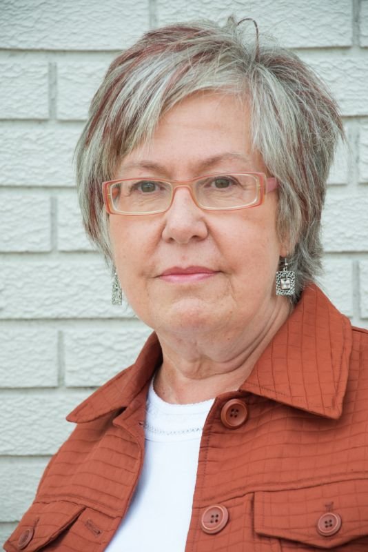 Portrait of an older woman wearing glasses, earrings, and a rust-colored jacket, standing in front of a white brick wall.