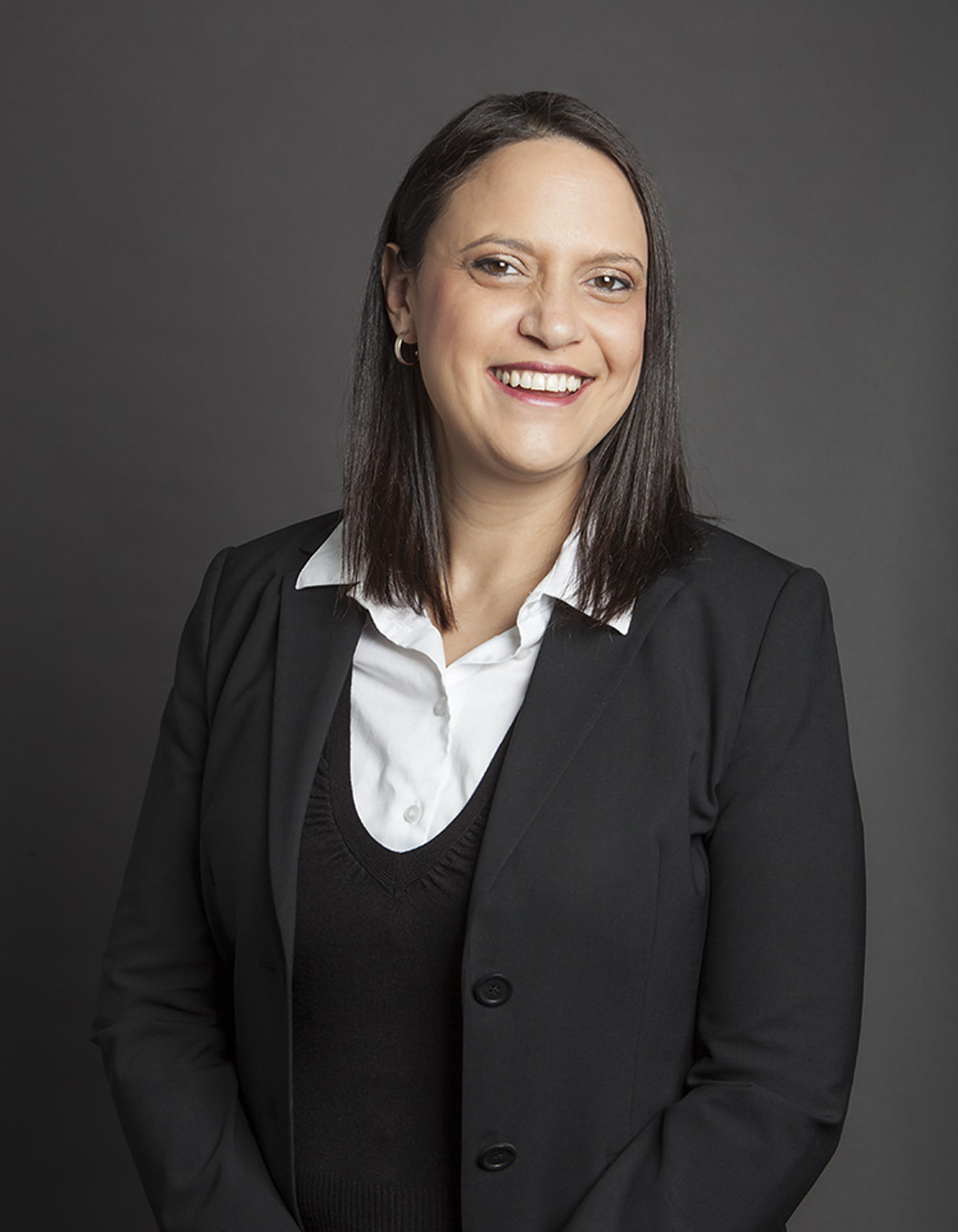 Professional portrait of a woman with dark brown hair, wearing a black blazer and white shirt, smiling against a gray background.