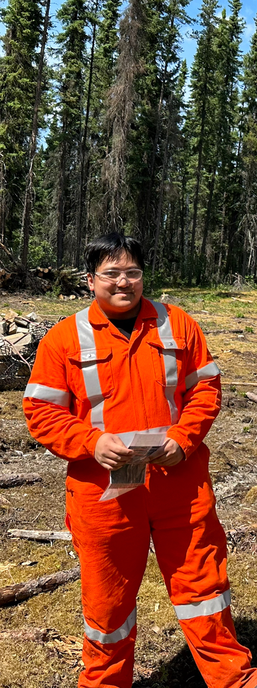 A person dressed in orange safety gear with reflective stripes standing outdoors in a forested area with tall trees and clear sky.