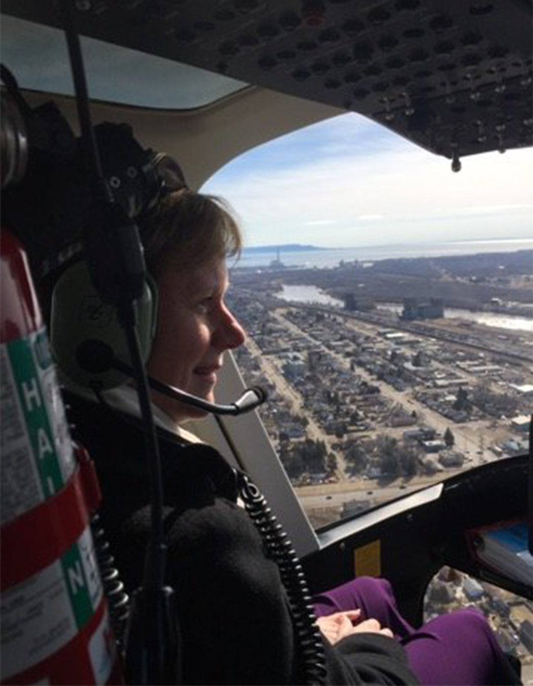 A woman is flying in a helicopter, looking out the window at the city and water below.