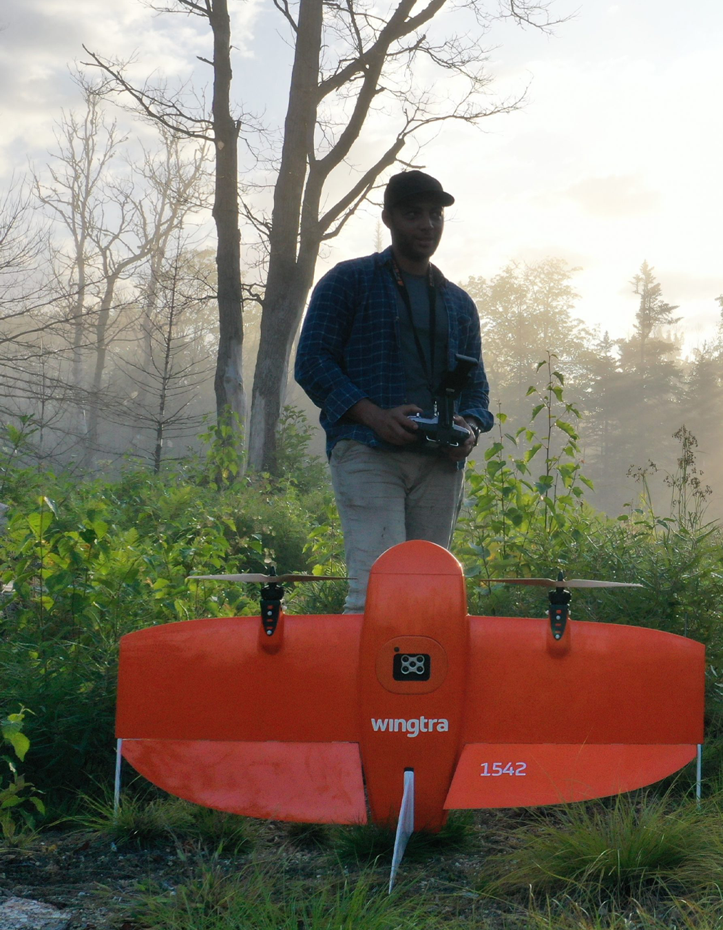 A man standing outdoors in a forested area with leafless trees in the background, holding a remote control. In front of him is a large, orange, drone-like aircraft with the brand name 'wingtra' and the number '1542' on it.