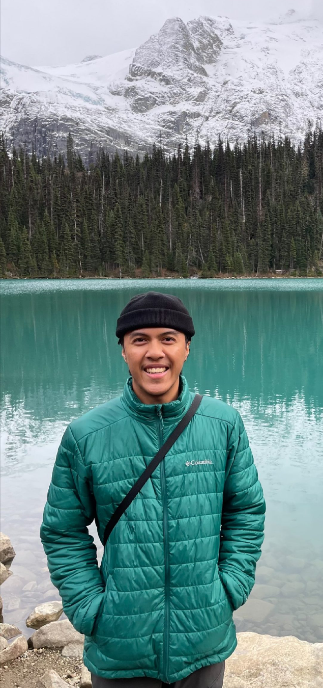 A smiling young man wearing a black beanie and green puffer jacket standing in front of a turquoise lake with a forested shoreline and snow-capped mountains in the background.