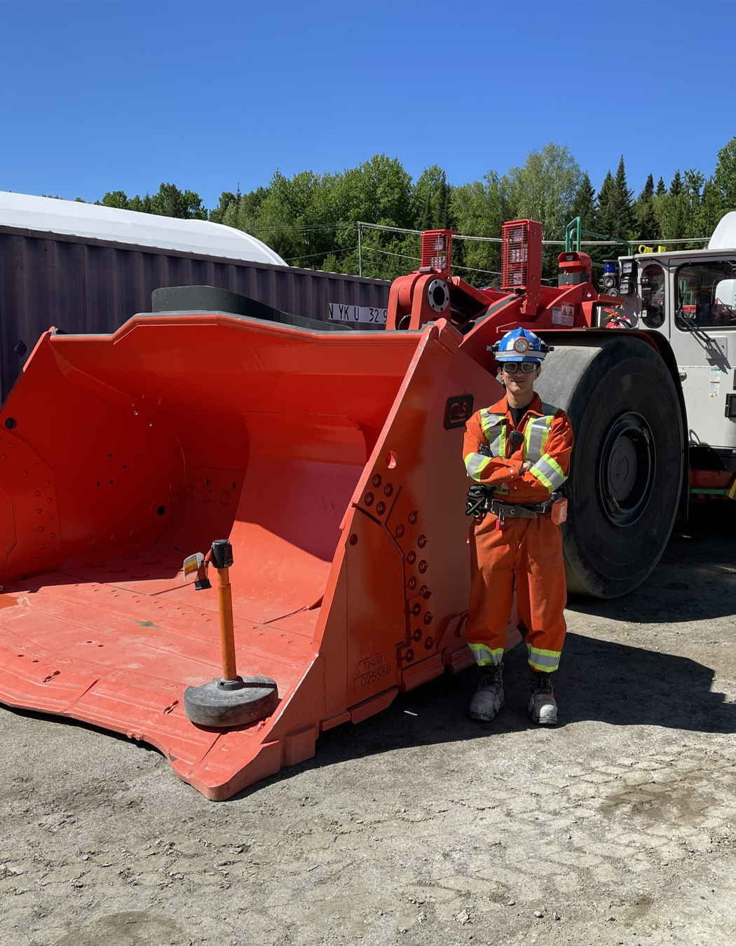 Construction worker in orange safety gear and helmet standing next to a large snowplow blade attached to a heavy-duty vehicle on a construction site. Trees and a blue sky are in the background.