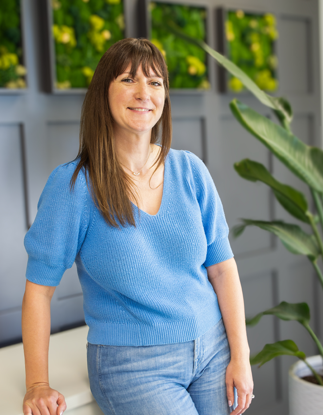 A woman with brown hair and a blue knitted top smiling and standing indoors with green plants in the background.