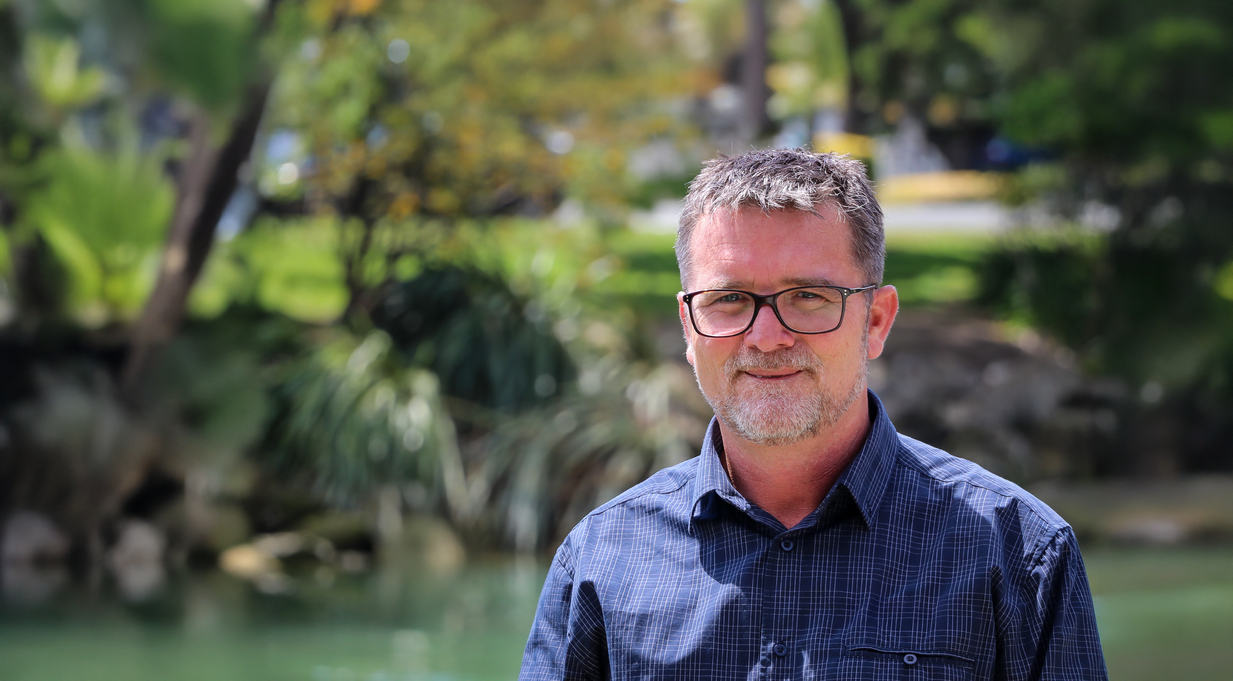 Nigel Skeggs, standing for Nelson City Council, standing outdoors near a body of water with trees and greenery in the background.