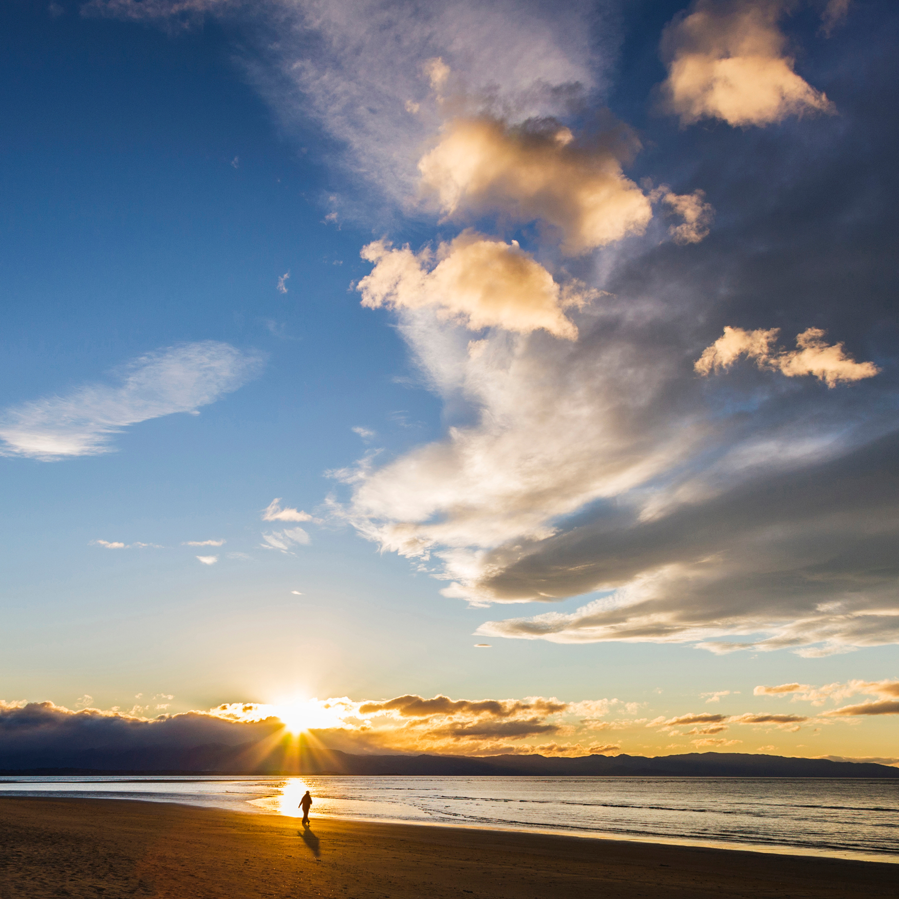 Sunset over the ocean with a person walking on the beach, clouds in the sky, and mountains in the distance.