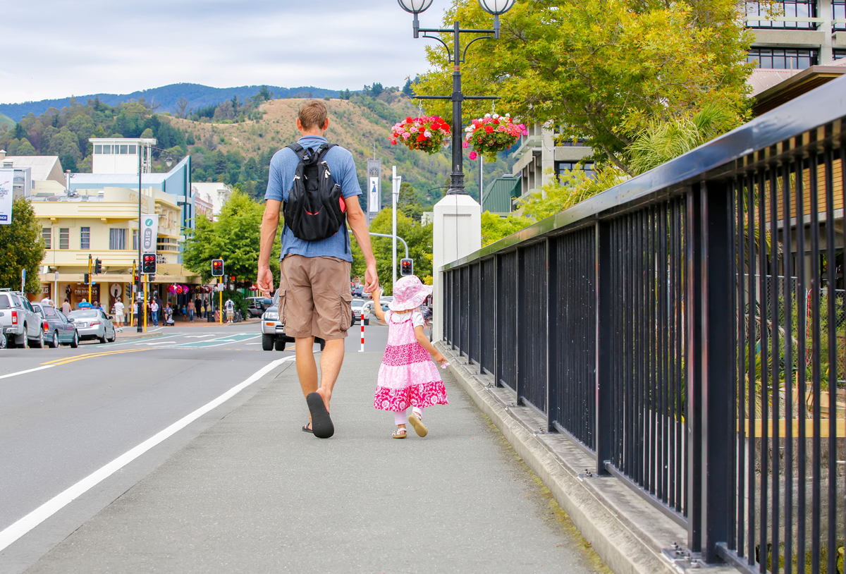 A man and a young girl walking on a city sidewalk, holding hands, with cars and buildings in the background, and a large tree and hanging flower baskets nearby.