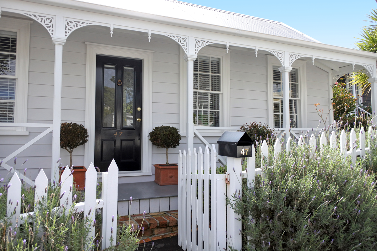 A white house with decorative trim, black front door, and a porch with white railings. There are two potted bushes on either side of the door, a black mailbox with the number 47, and a white picket fence with a small gate. Lavender plants are in the garden in front of the house.