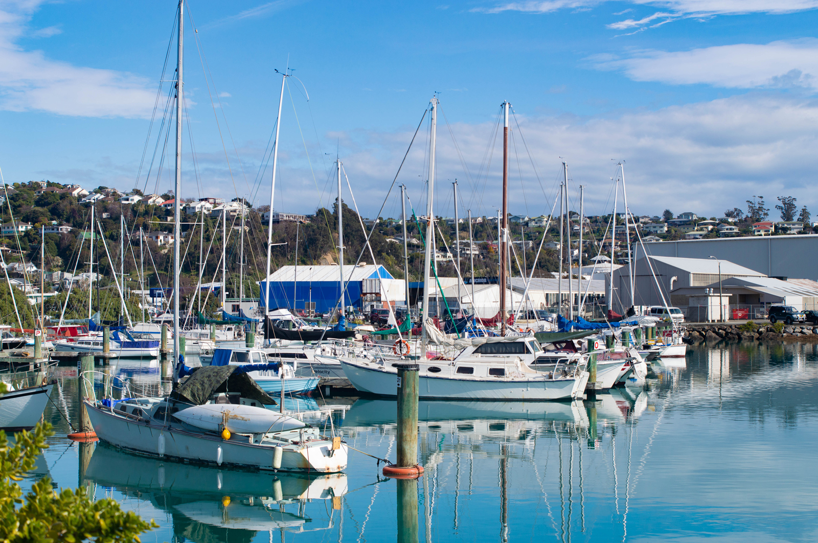 Marina with sailboats and yachts docked, buildings on hillside, partly cloudy sky.