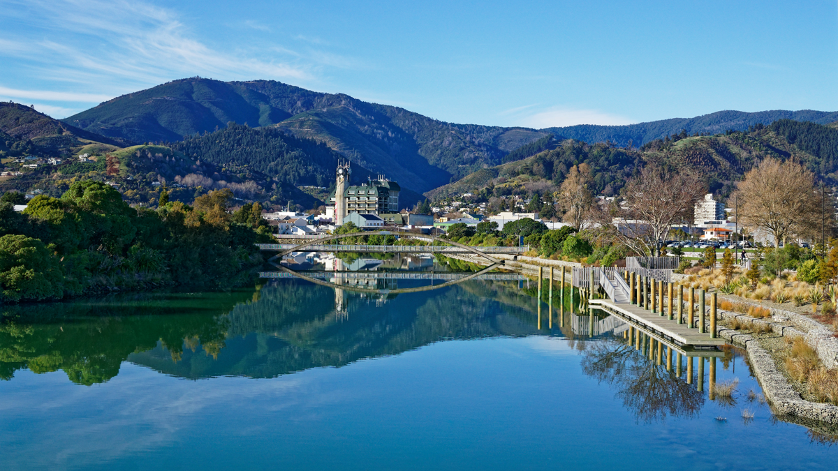 A peaceful river reflects the surrounding trees and buildings in a small town set against green mountains under a clear blue sky.