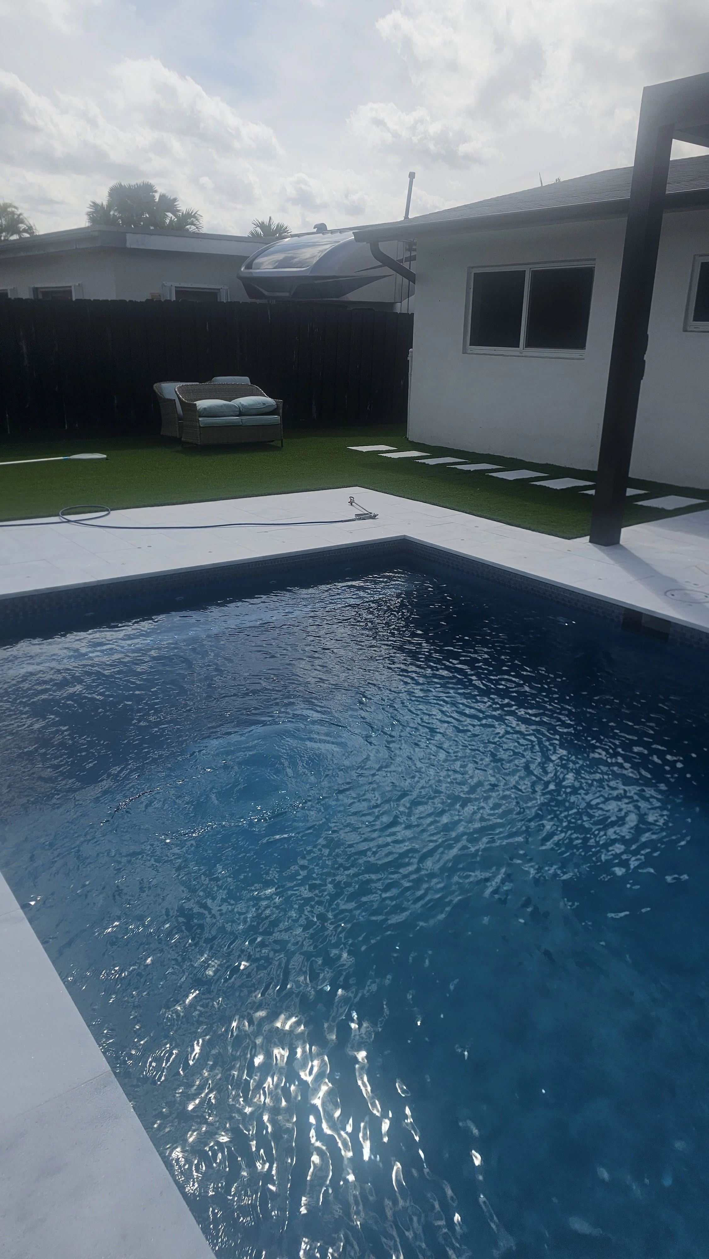 A backyard with a swimming pool, a small grassy area, a two-seater outdoor sofa, a black fence, an overcast sky, and a house with a window and a protruding awning.