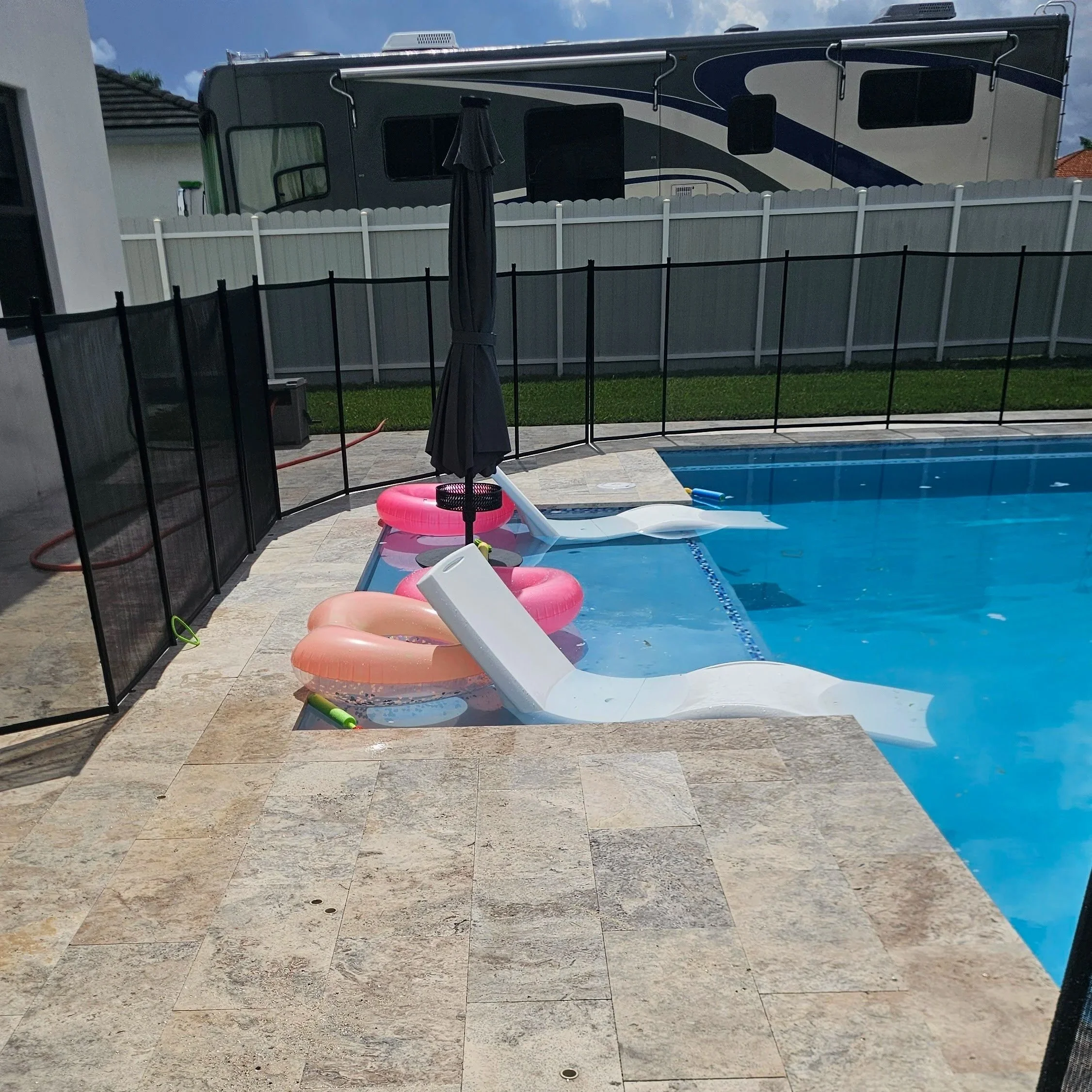 A swimming pool with three pool floats and a closed black umbrella, enclosed by black safety fencing, with a large RV parked in the background.