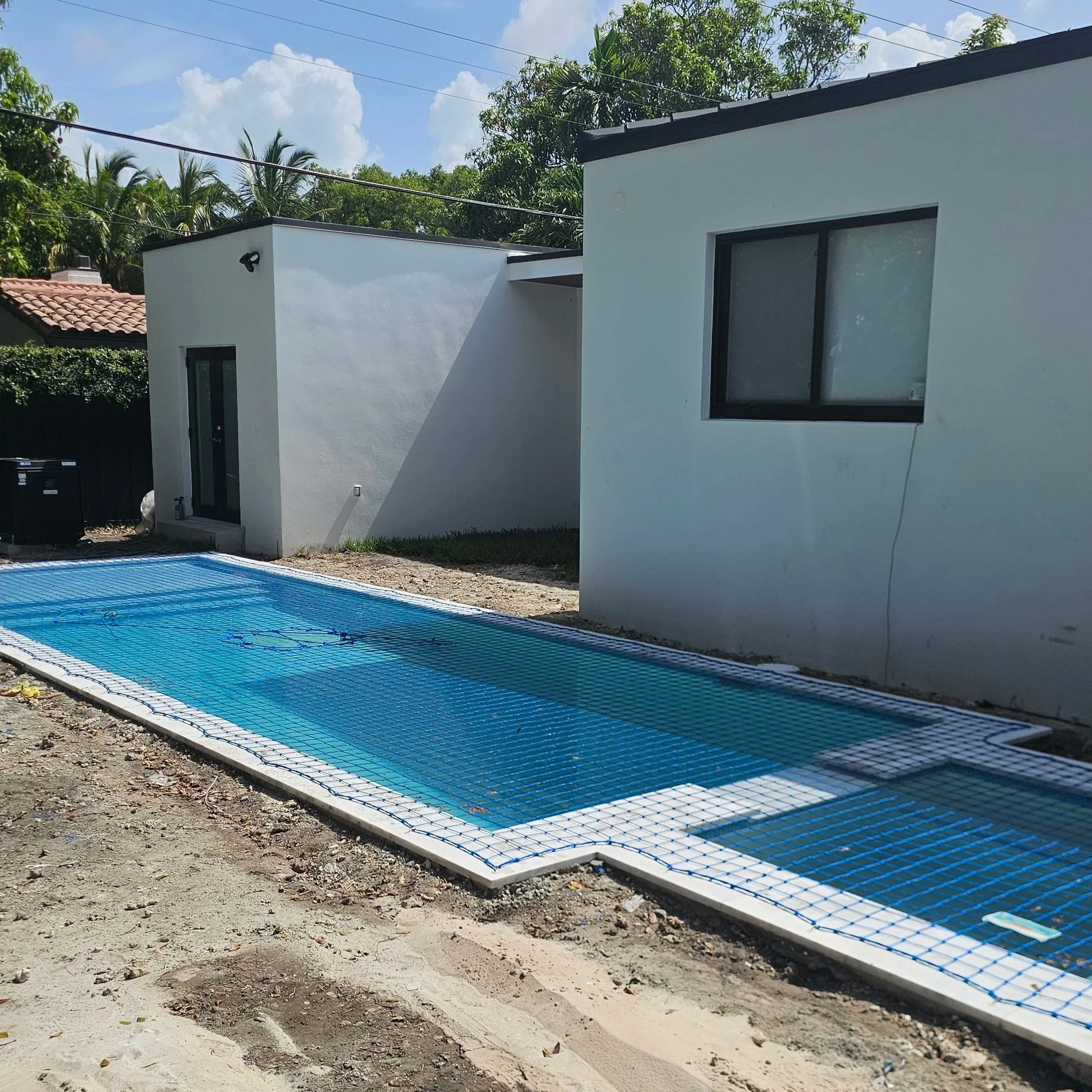 A modern backyard with a rectangular in-ground swimming pool covered with a safety net, adjacent to a white house with black-framed windows, and surrounded by trees and a partly cloudy sky.
