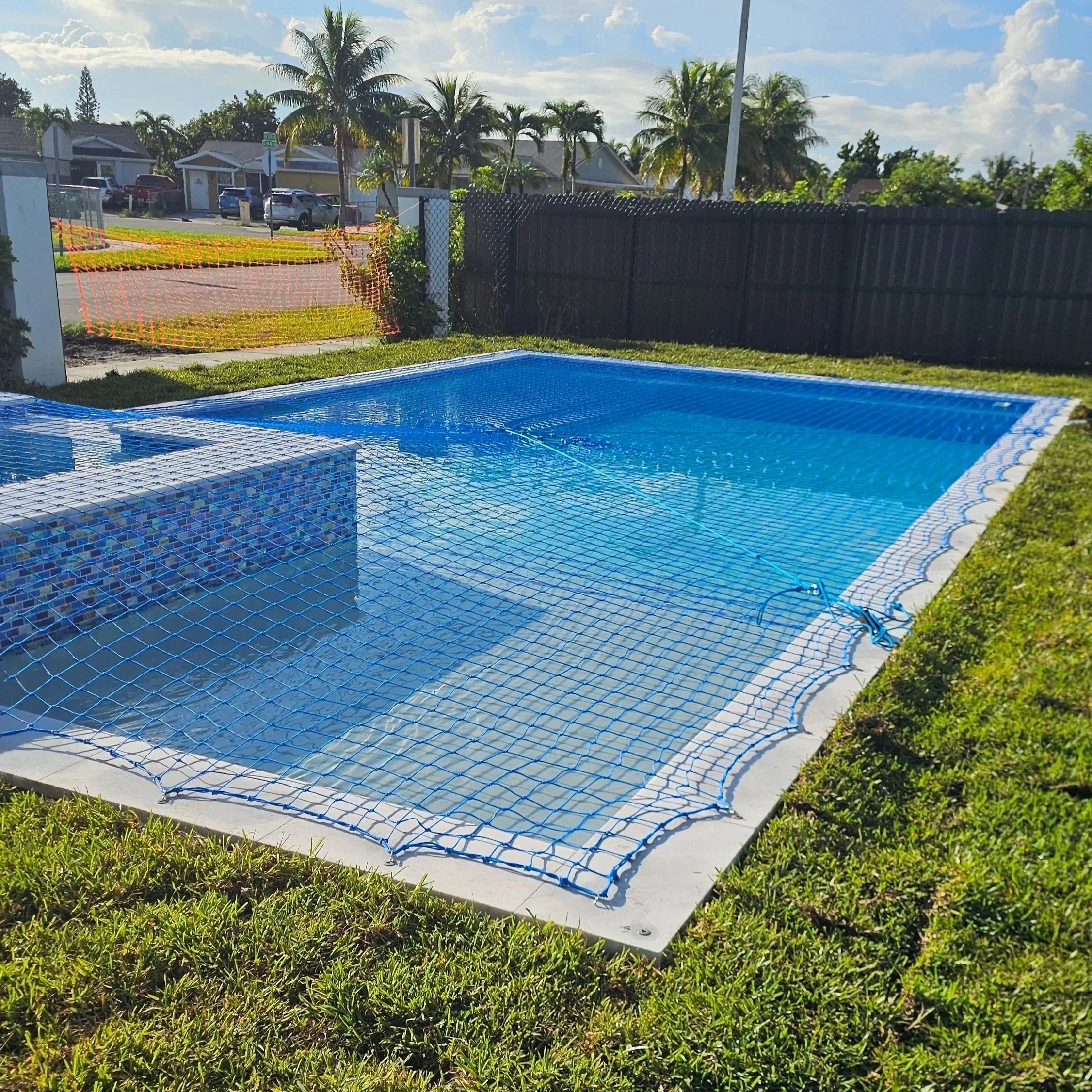 Swimming pool with a safety net overhead, surrounded by grass, in a backyard with a house, trees, and parked cars visible in the background.