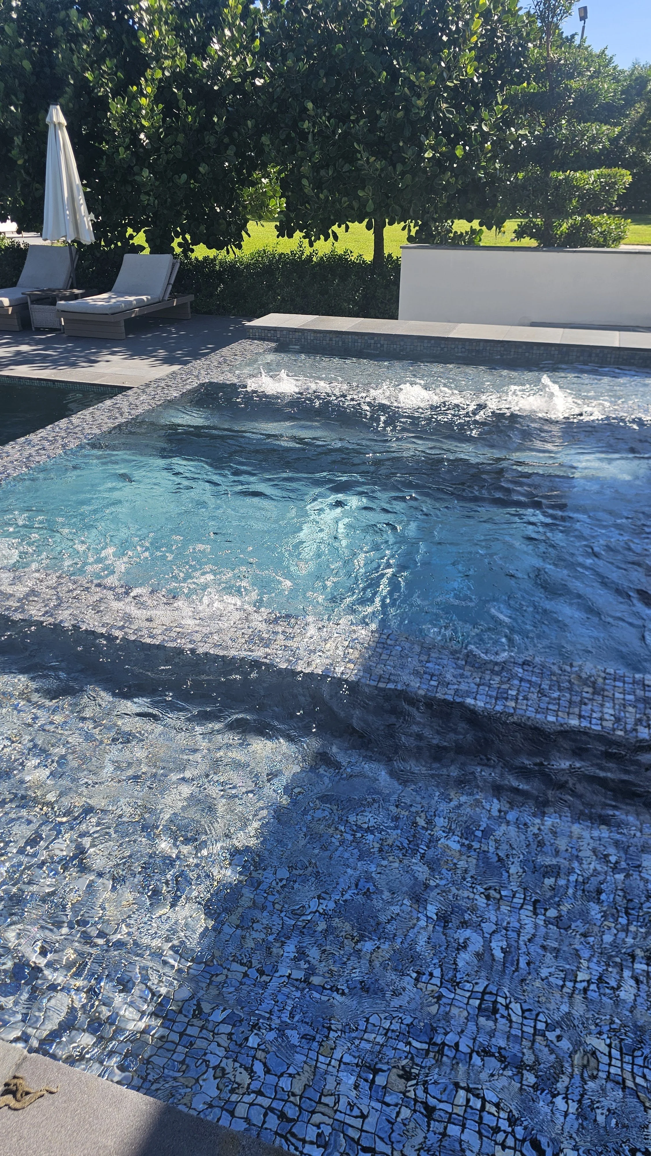 A backyard swimming pool with a side hot tub, lounge chairs, a closed white umbrella, and lush green trees and bushes in the background.