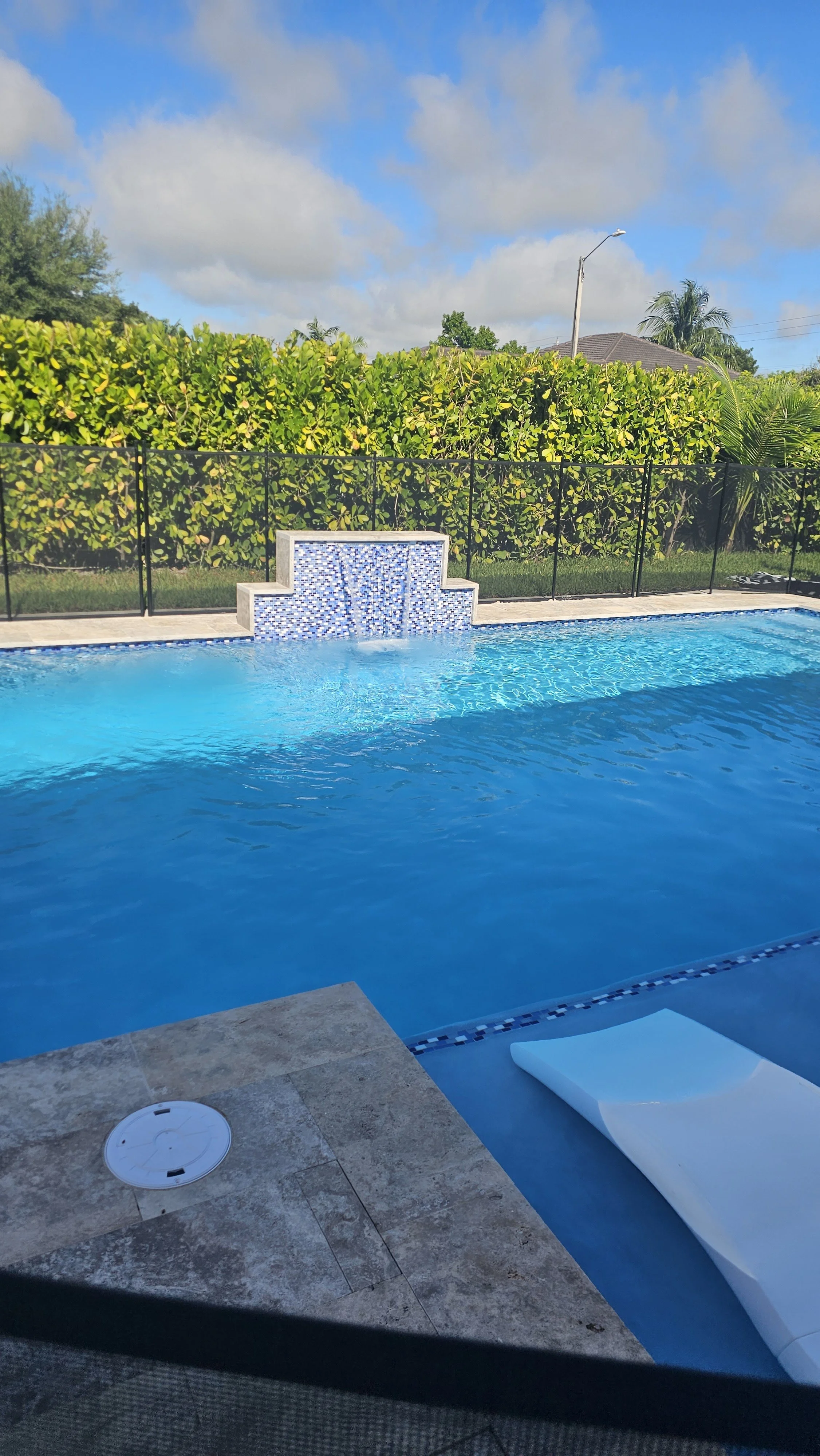 A backyard swimming pool with blue water, surrounded by a stone deck, with a waterfall feature, a black safety fence, green hedge, trees, a lawn, a house, and a lamppost under a partly cloudy sky.