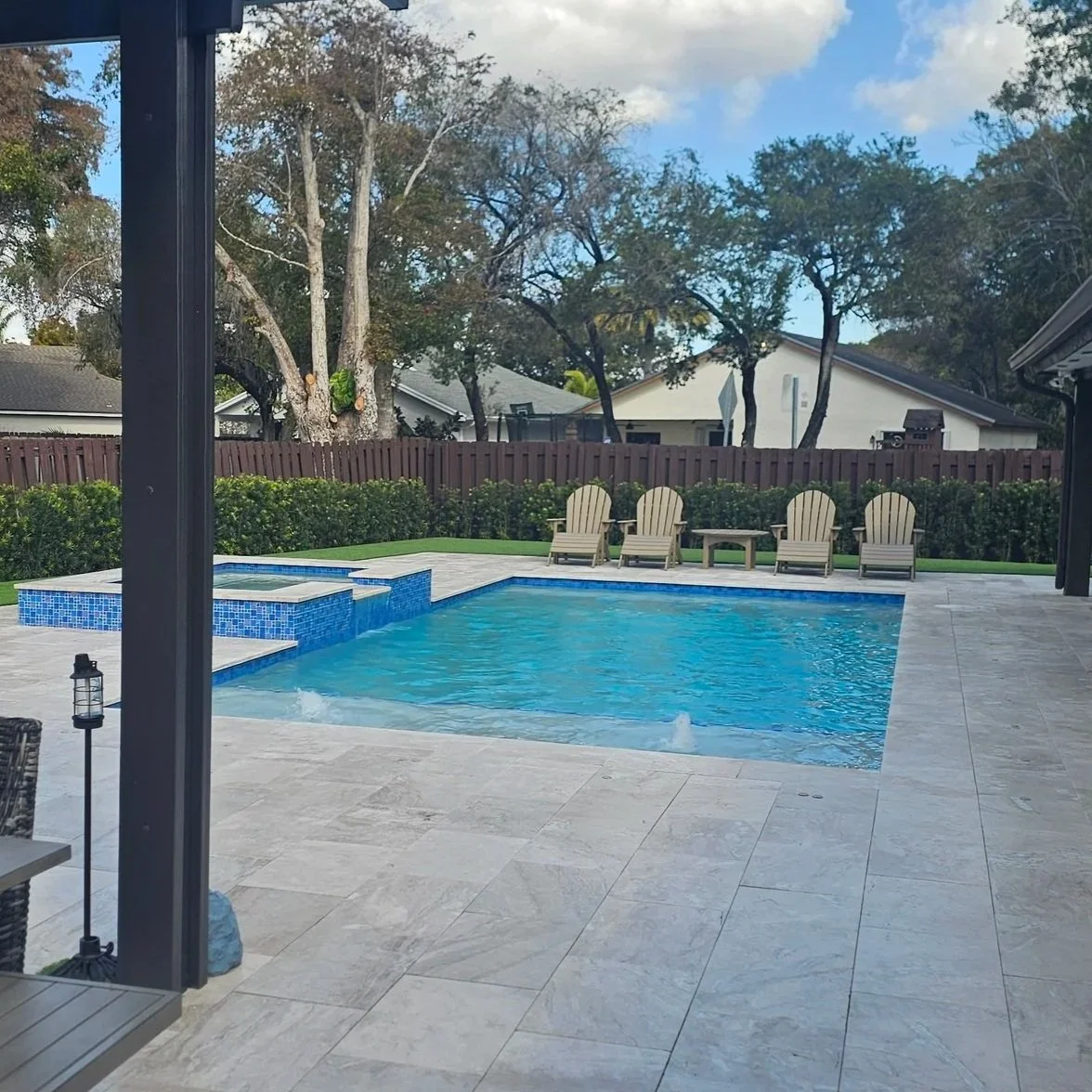 A backyard with a swimming pool featuring a hot tub and five Adirondack chairs facing the pool, surrounded by greenery and trees, with a wooden fence and houses in the background.