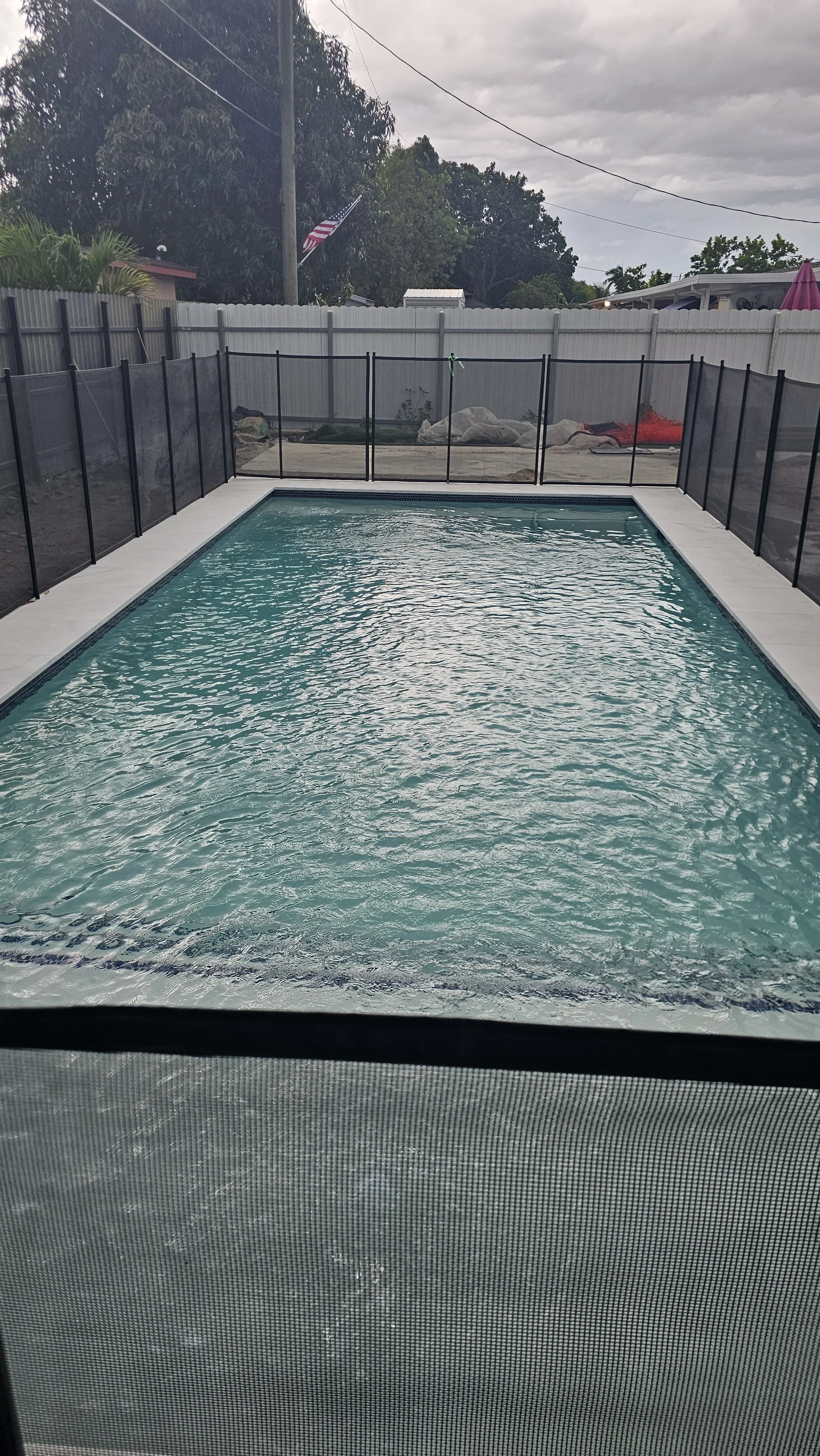A rectangular swimming pool with a black safety fence around it, set in a backyard with trees, a fence, and some outdoor furniture in the background, under a cloudy sky.