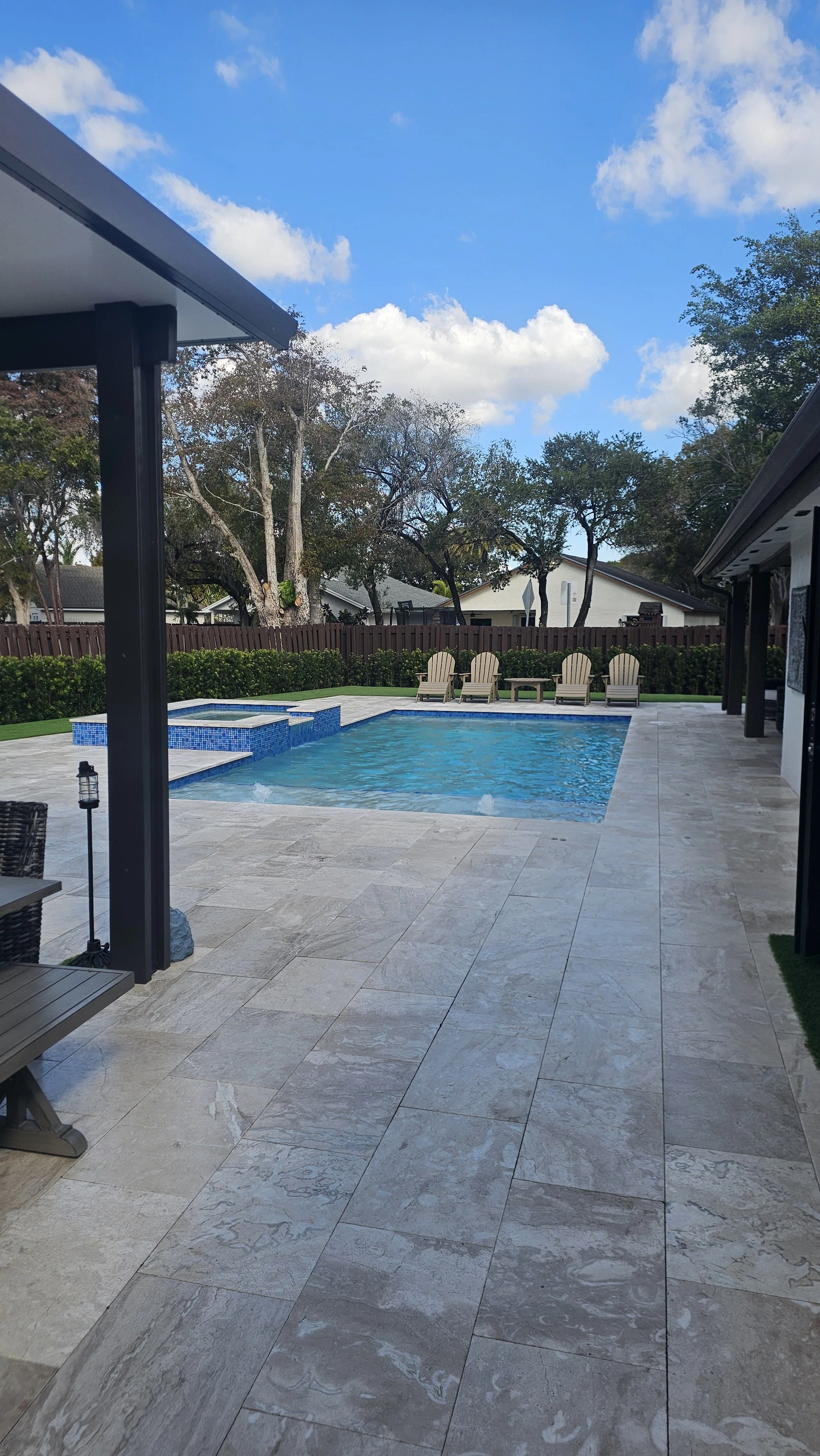 View of a backyard pool with four Adirondack chairs and a wooden fence, trees, and a partly cloudy sky in the background.