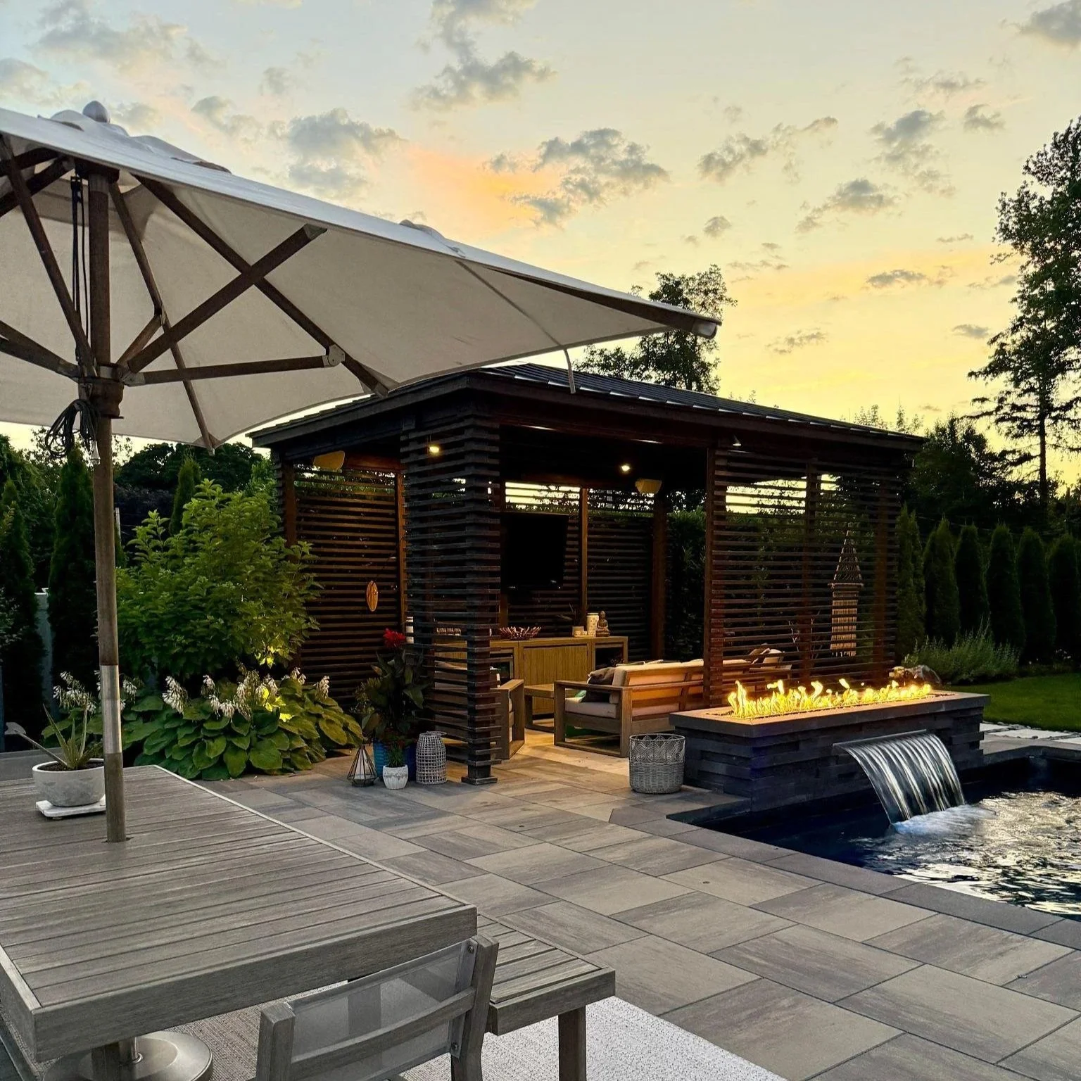 A backyard patio featuring a pool with a waterfall, a fire feature, a shaded sitting area with outdoor furniture, a large umbrella, and lush greenery, during sunset.