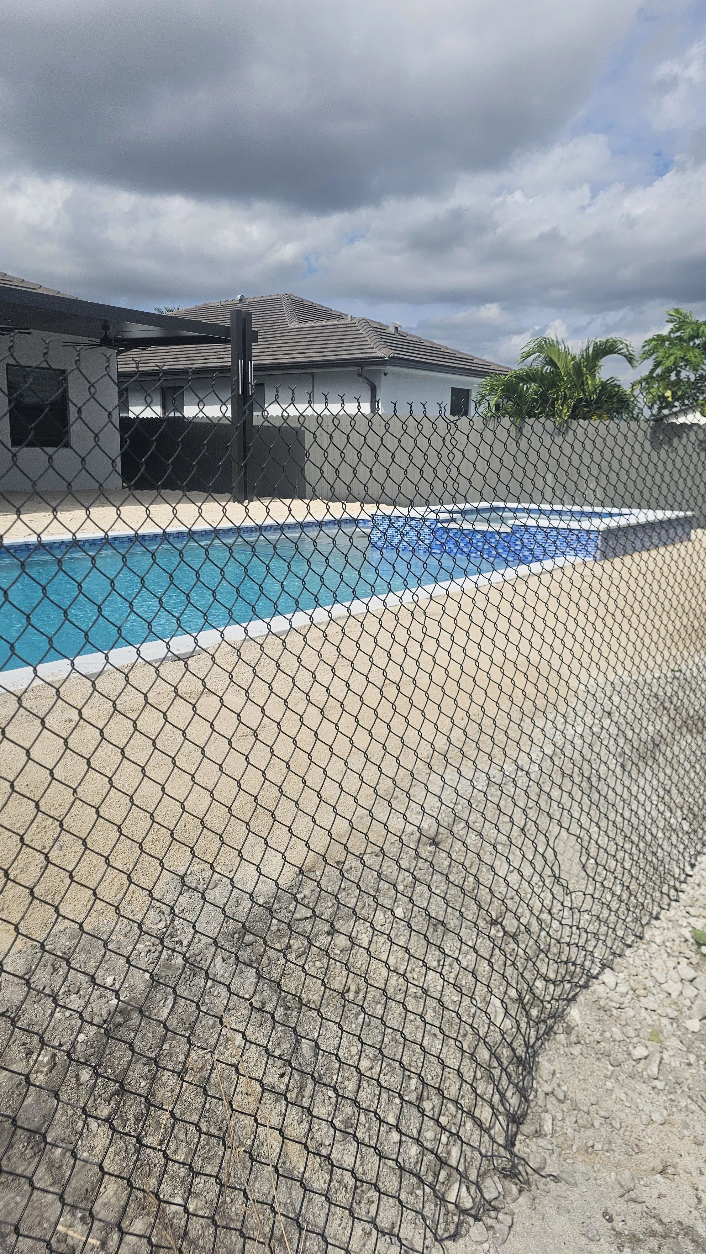 A backyard with a swimming pool surrounded by a black chain-link fence, a beige concrete deck, a gray house with a tiled roof, a tree with green leaves, and a cloudy sky.