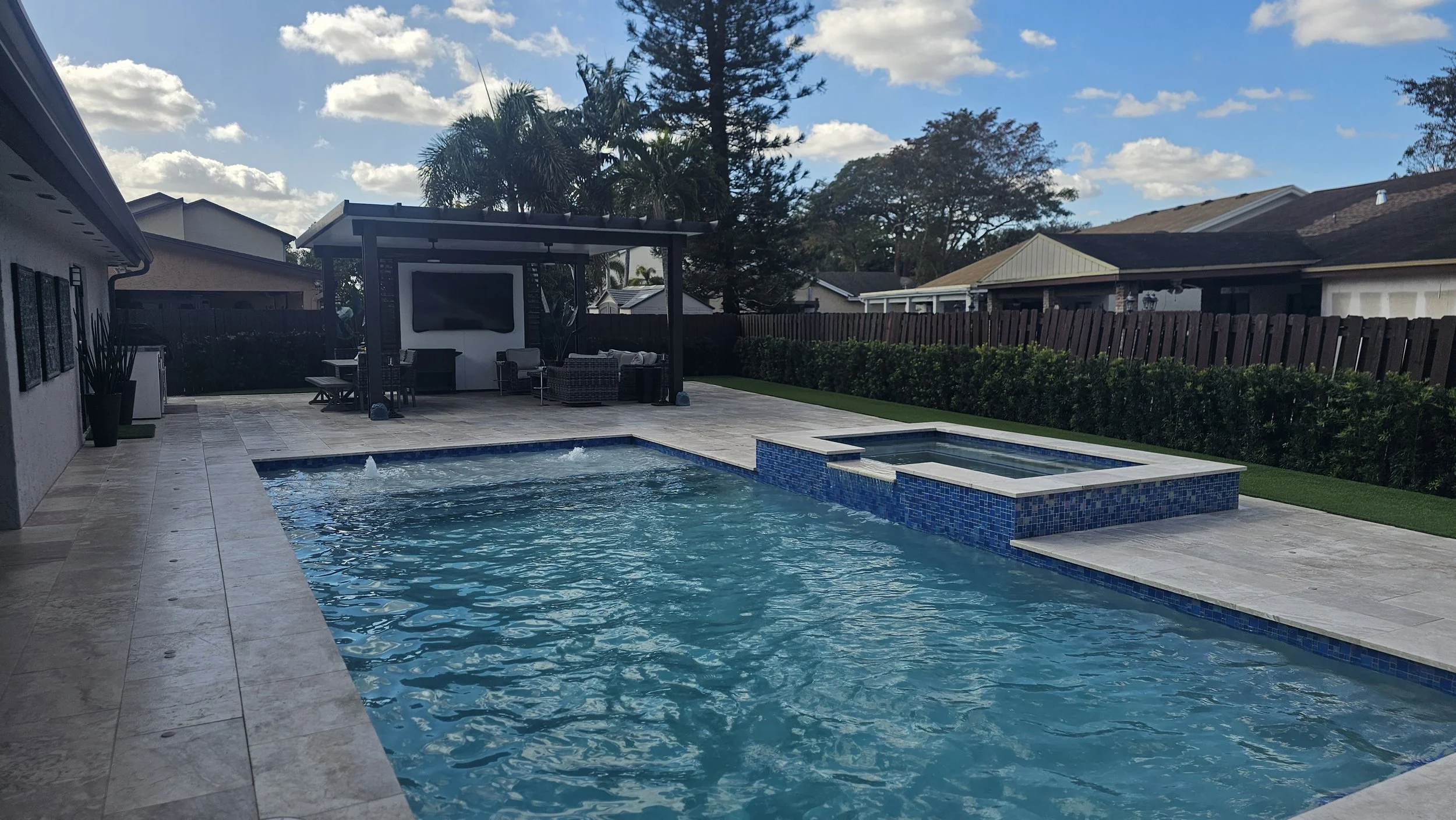 Backyard swimming pool with a hot tub, surrounded by a tiled patio, with outdoor furniture and a screened TV area, fenced yard with trees and bushes.