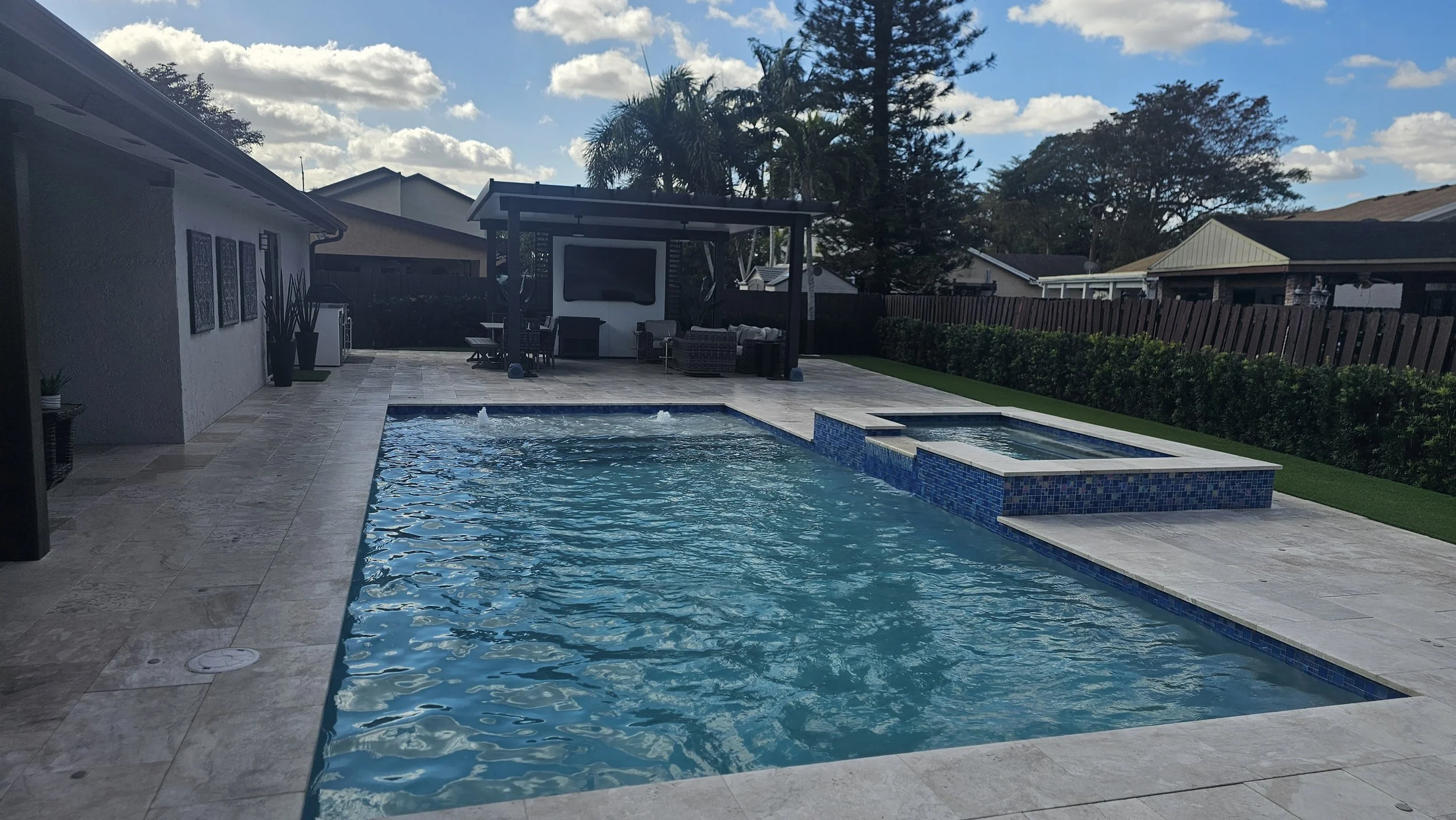 Backyard with rectangular swimming pool and attached hot tub, surrounded by a beige stone patio, with outdoor furniture and a shaded seating area, fenced yard, and some trees and houses in the background under a partly cloudy sky.