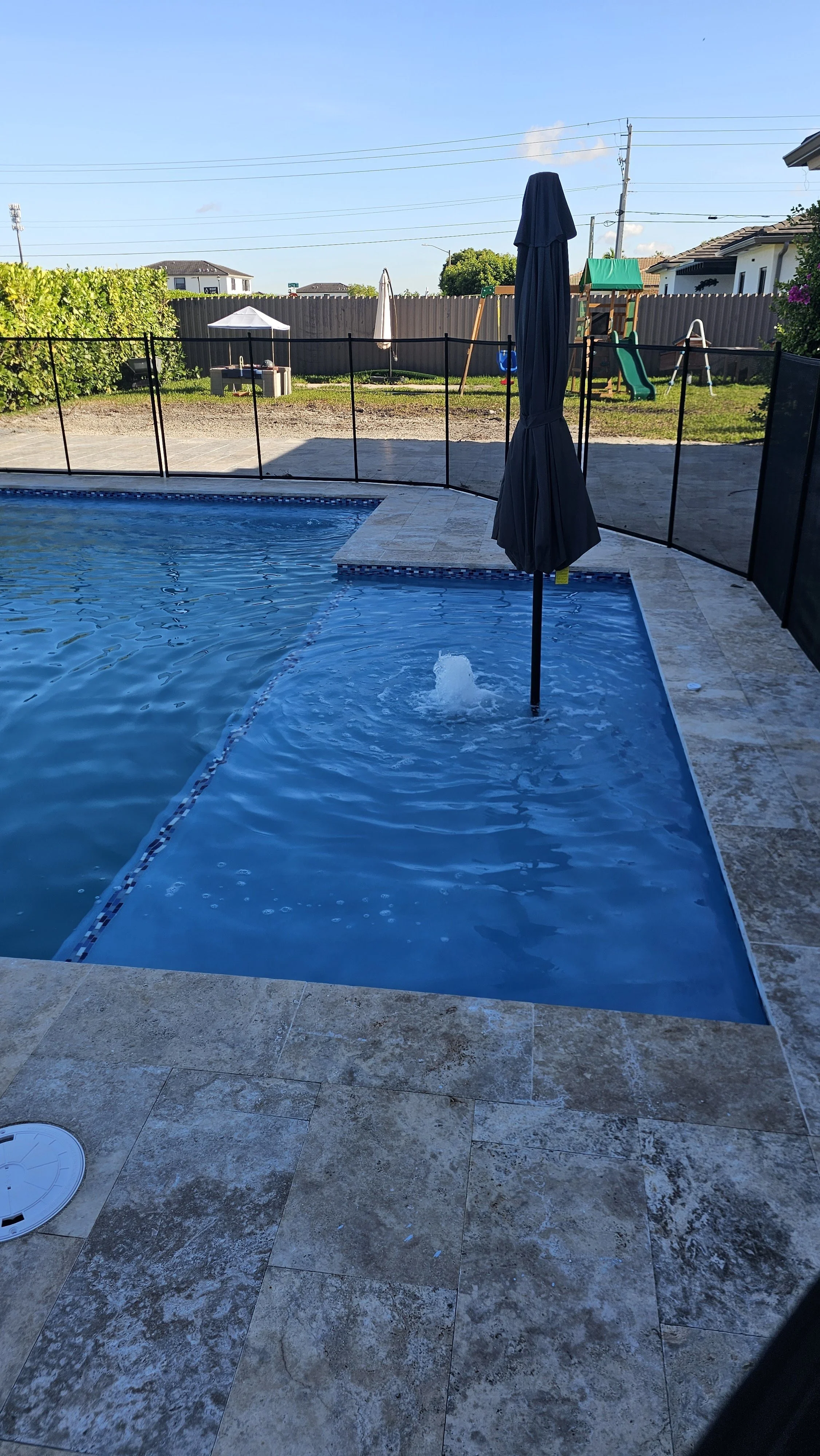 A swimming pool with a closed dark-colored umbrella in the water, surrounded by tiled pool deck, with a backyard playground and a fence in the background, under a clear blue sky.