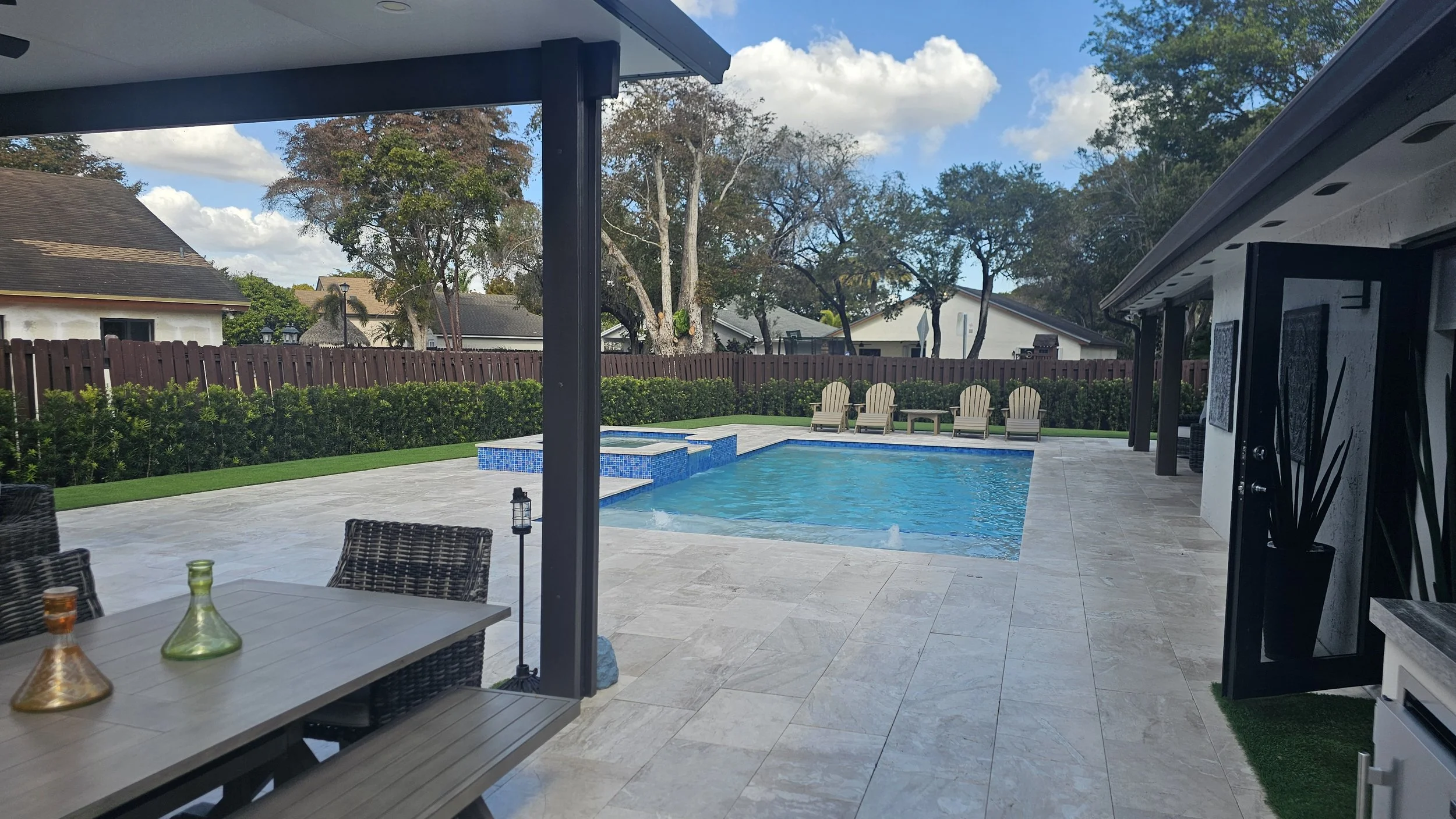 A backyard with a swimming pool, patio furniture, and lush green trees and bushes. There are lounge chairs on the far side of the pool and a fenced yard with houses and trees in the background.