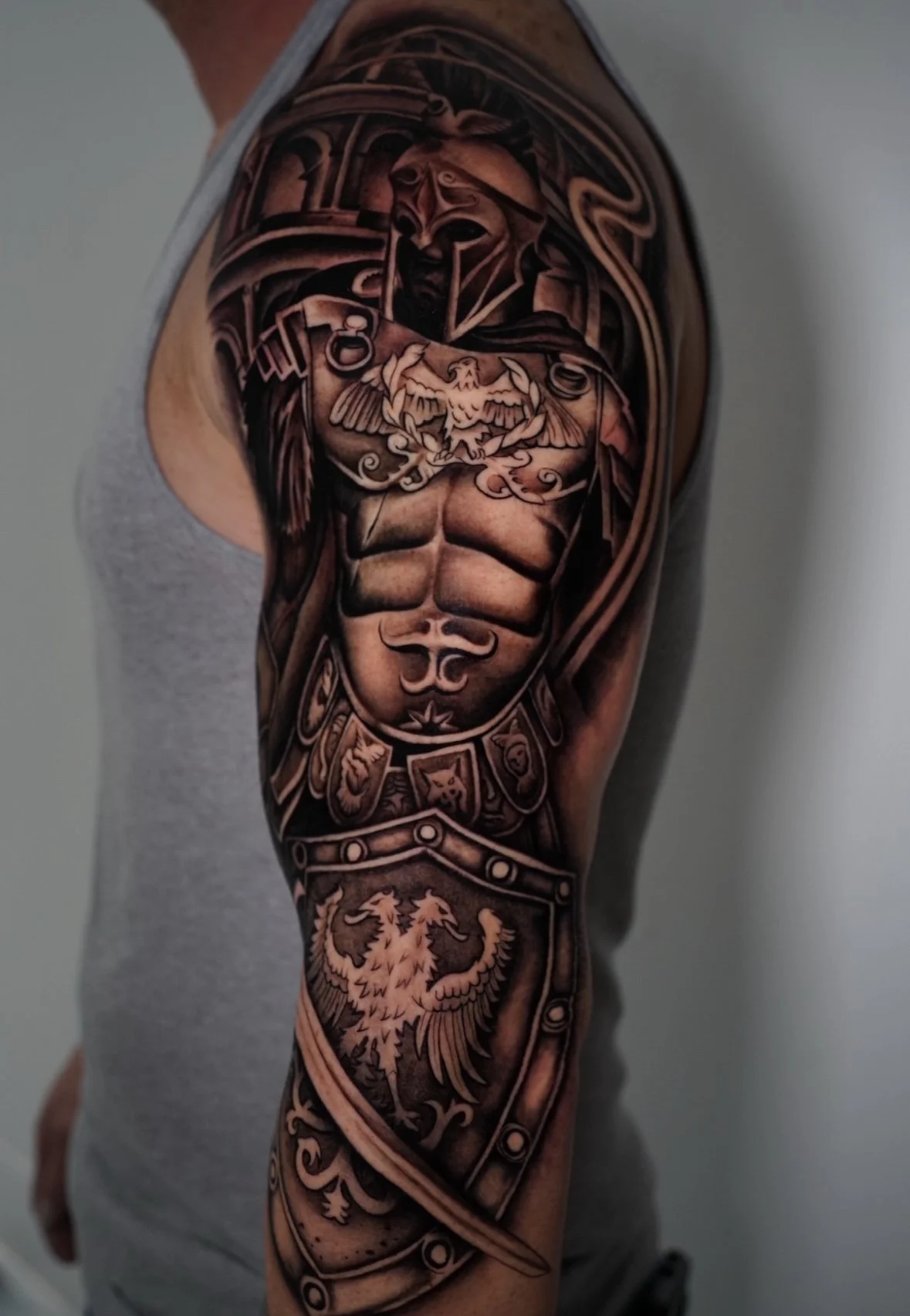 A detailed black and gray tattoo of a Roman soldier with armor, a helmet, facial beard, and a shield with a lion emblem on the man's upper arm.