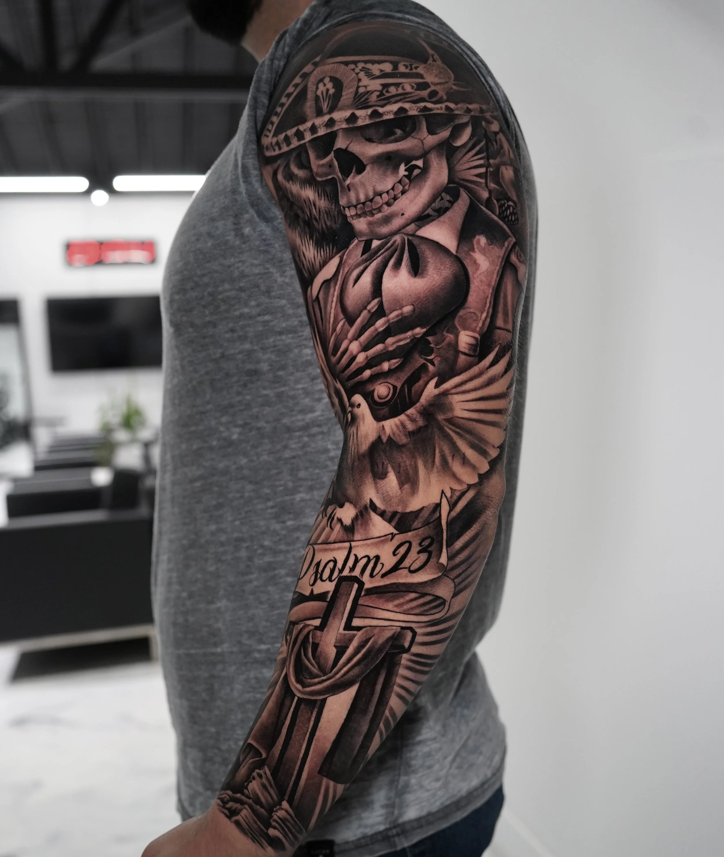 Black and gray tattoo sleeve featuring a skeleton with a skull and army helmet, a dove, a cross with a ribbon reading "Ojam 23," and various shading details.