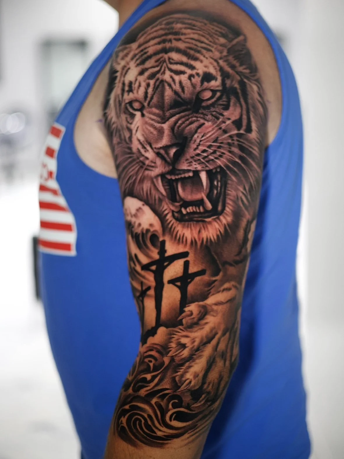Tattoo of a roaring tiger's face on man's upper arm with crosses and swirling patterns.