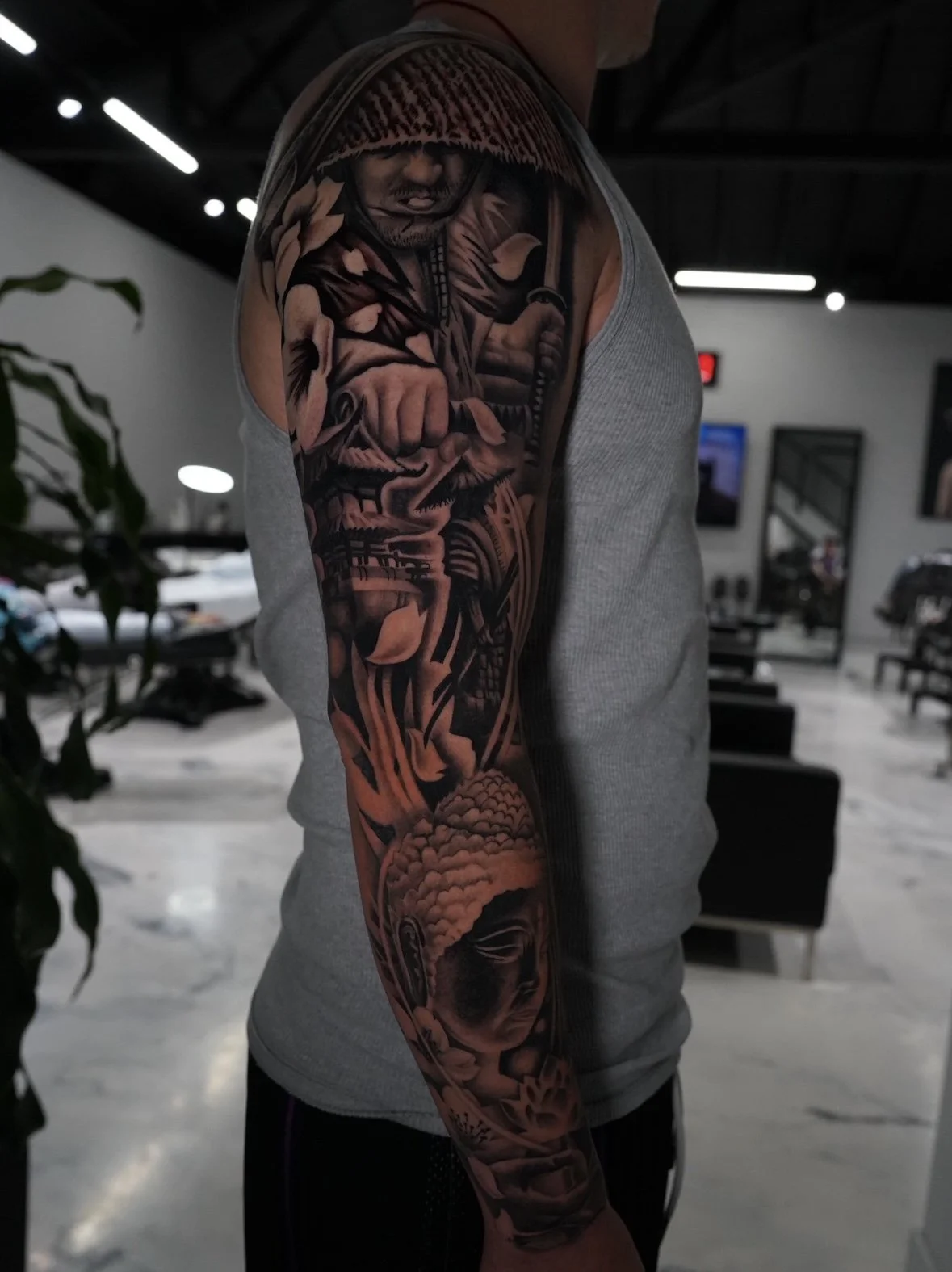 Close-up of a person's arm with detailed black and gray tattoo artwork, featuring a man holding a sword and a Buddha head.