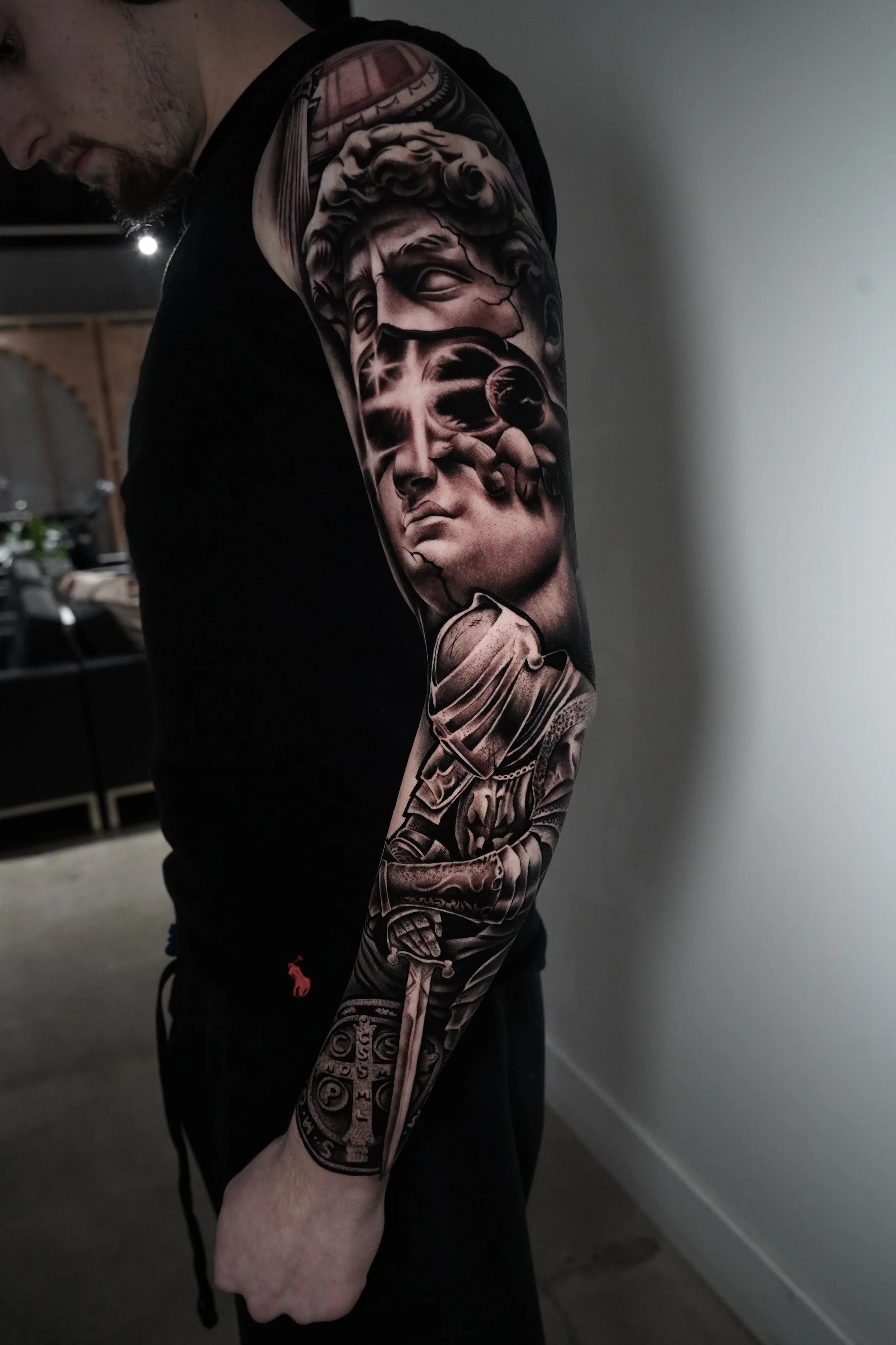 Black and gray sleeve tattoo featuring classical sculptures, a futuristic planet, and a knight with a sword.