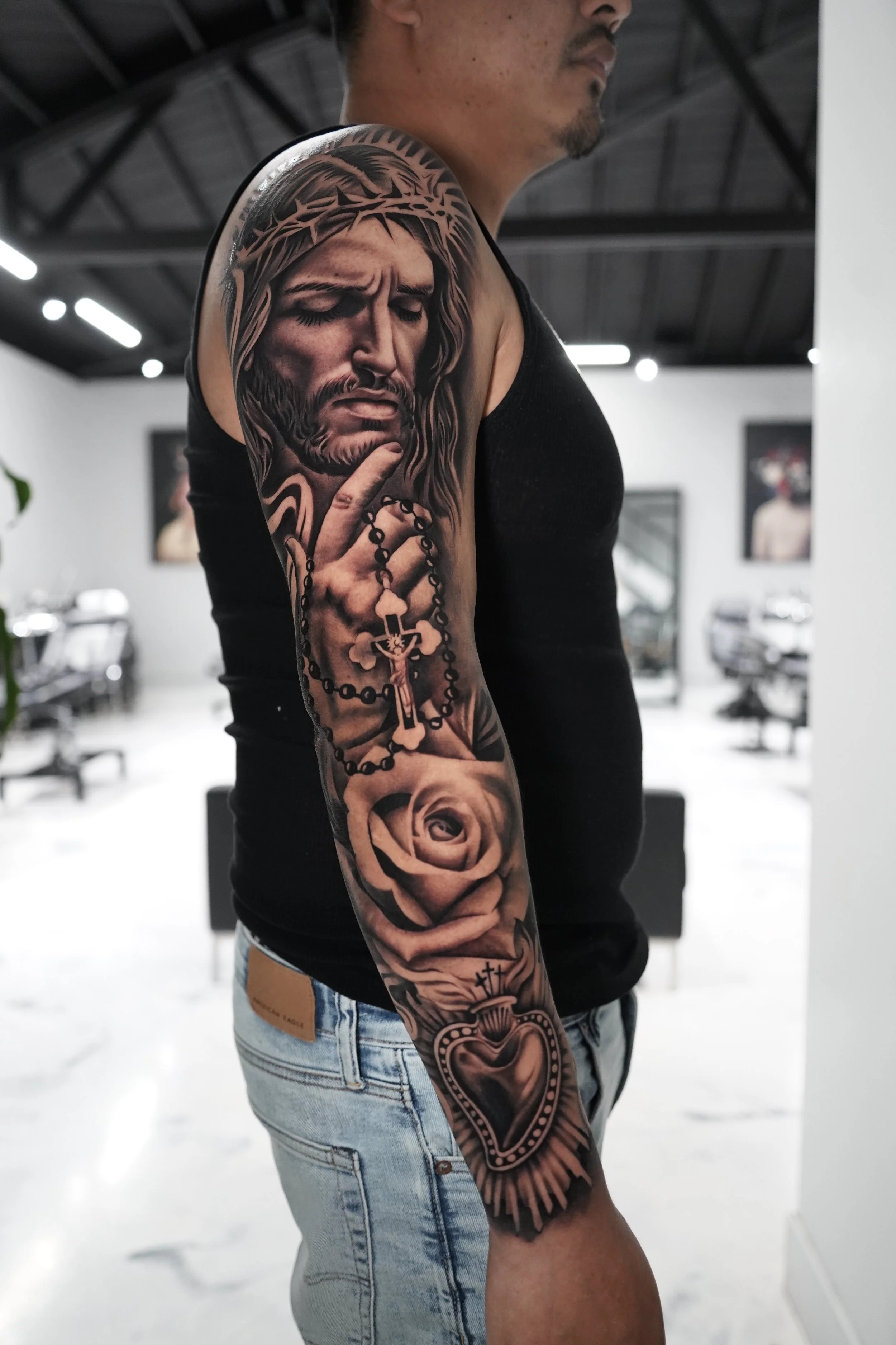 A person with a sleeve tattoo on their left arm featuring a realistic depiction of Jesus Christ with closed eyes, a crown of thorns, holding a rosary with a cross and a rose at the bottom, in black and gray ink.