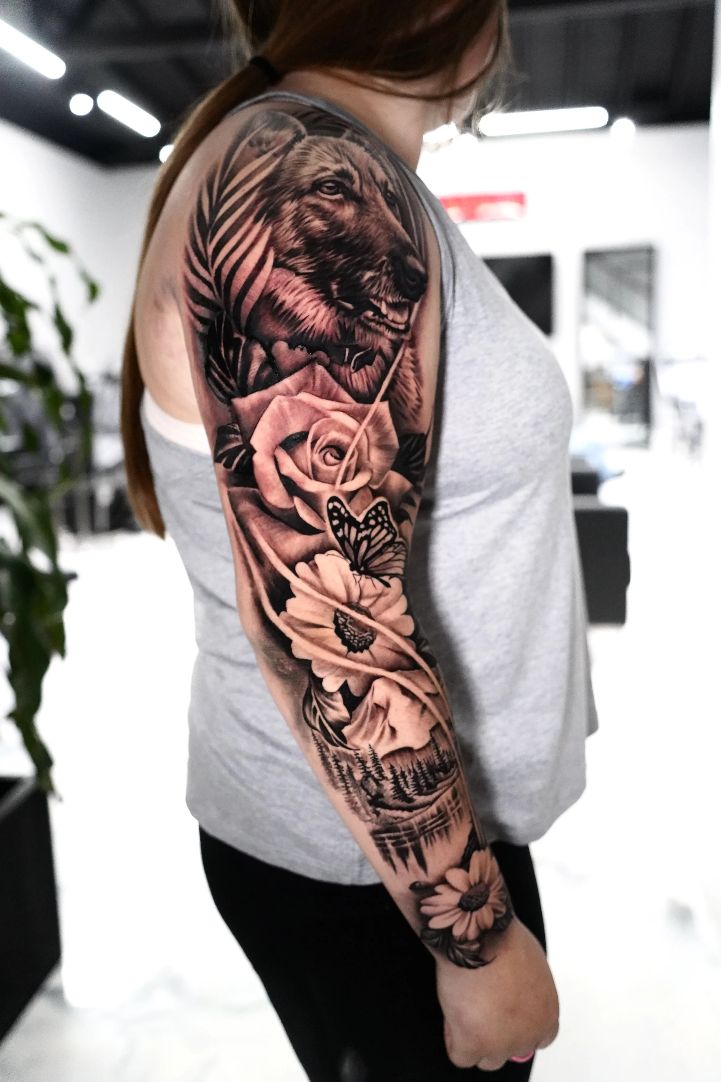 Tattoo of a bear's head, roses, butterflies, and a mountain landscape on a woman's upper arm.