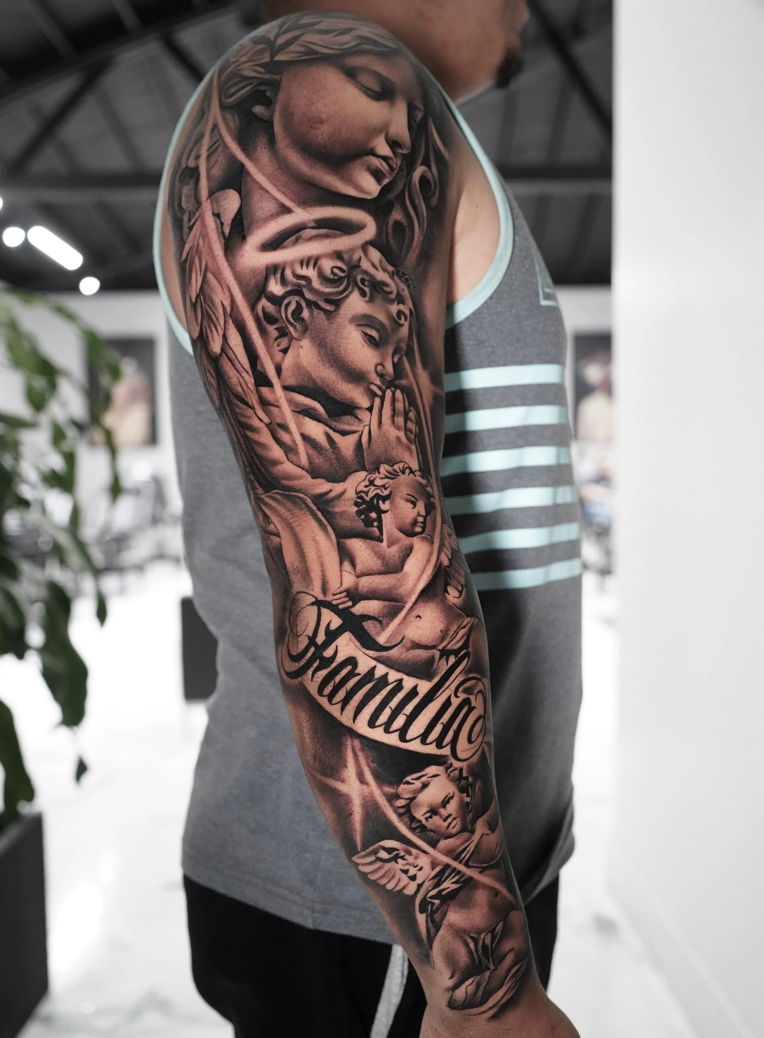 Close-up of a person's upper arm with detailed black and gray tattoo featuring religious figures, angels, and the word 'Familia' in script.