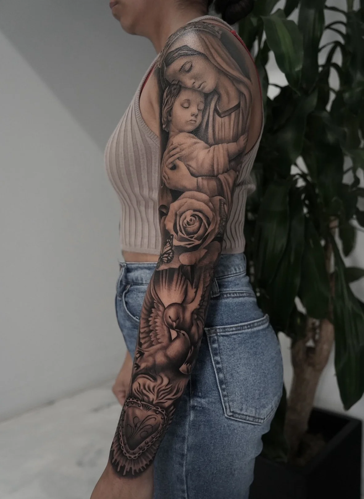 A person with a detailed black and gray tattoo sleeve featuring a mother and child, roses, a butterfly, a flying bird, and a heart.