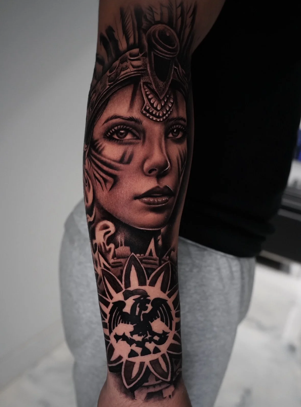 A detailed black and gray tattoo on a person's arm featuring a woman's face with tribal face paint, surrounded by tribal and symbolic elements.