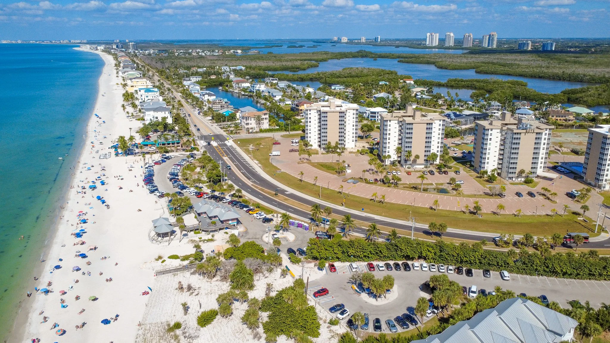 Aerial view of a beachside with white sandy beach, lined with colorful umbrellas, next to a row of beachfront structures, with a parking lot and high-rise buildings in the background, and water bodies with lush green surroundings.